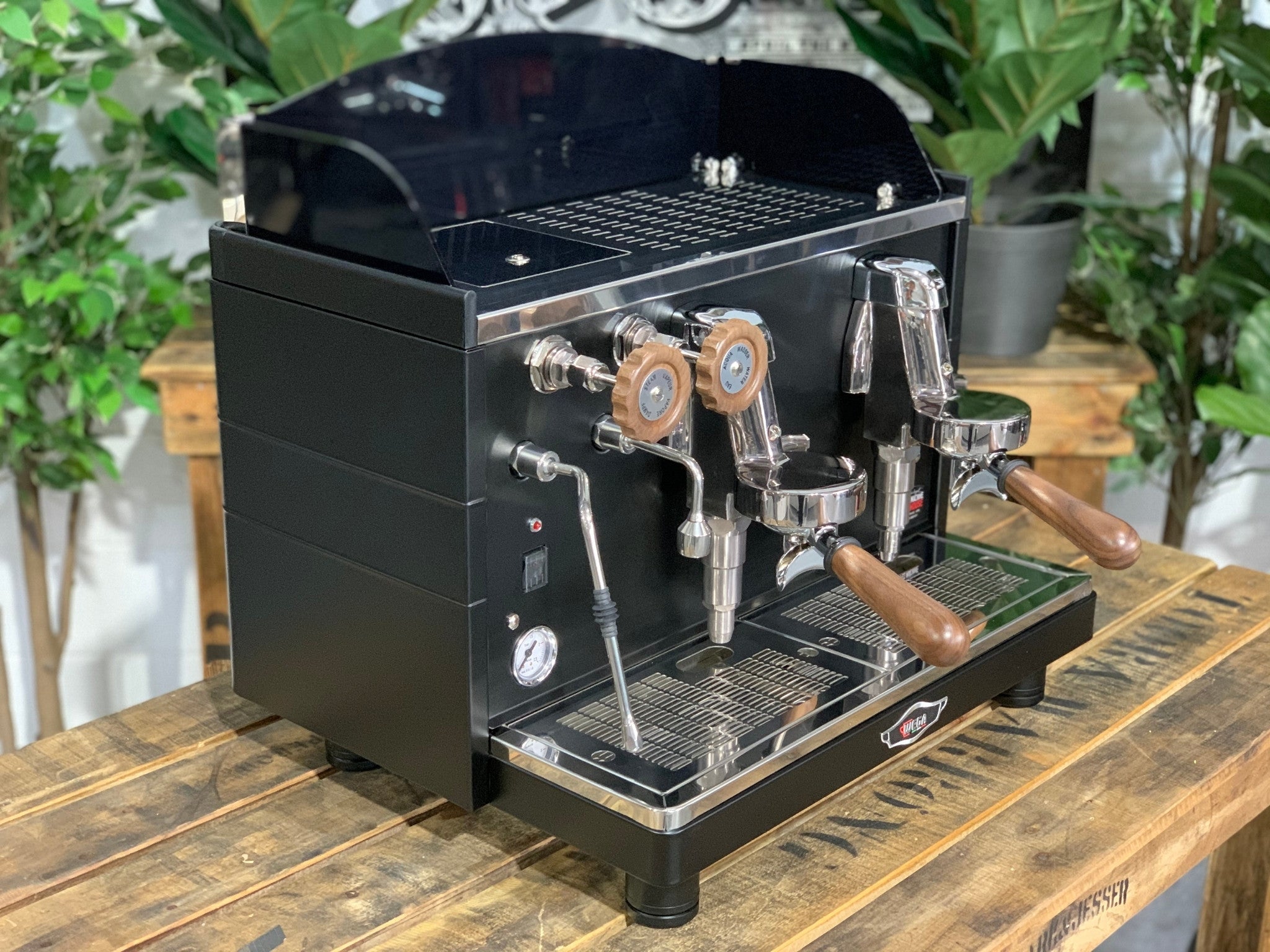 A brand new Wega Mini Nova Classic 2 Group Coffee Machine with a Matte Black finish & Timber Accents is sitting on a wooden bench. There is greenery in the background. The image is taken from the front of the machine and the machine is sitting on an angle so that you can see the front of the machine and the side.