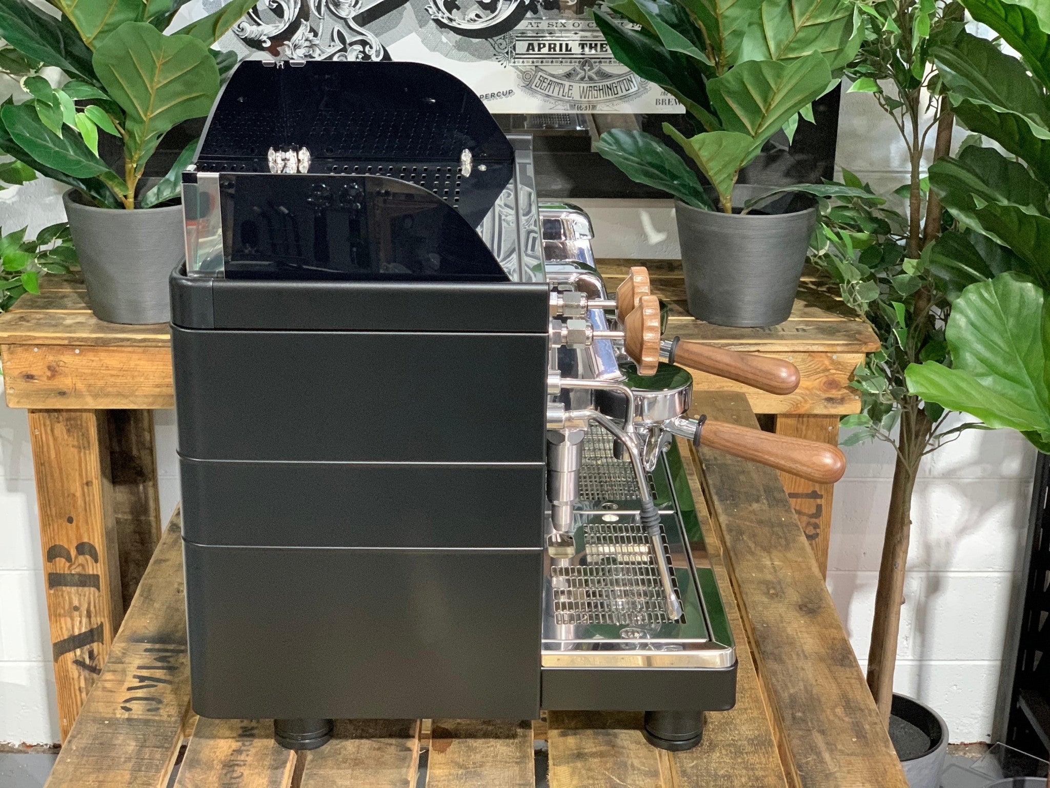 A brand new Wega Mini Nova Classic 2 Group Coffee Machine with a Matte Black finish & Timber Accents sitting on a wooden bench. There is greenery in the background. The image is taken from the side of the machine.