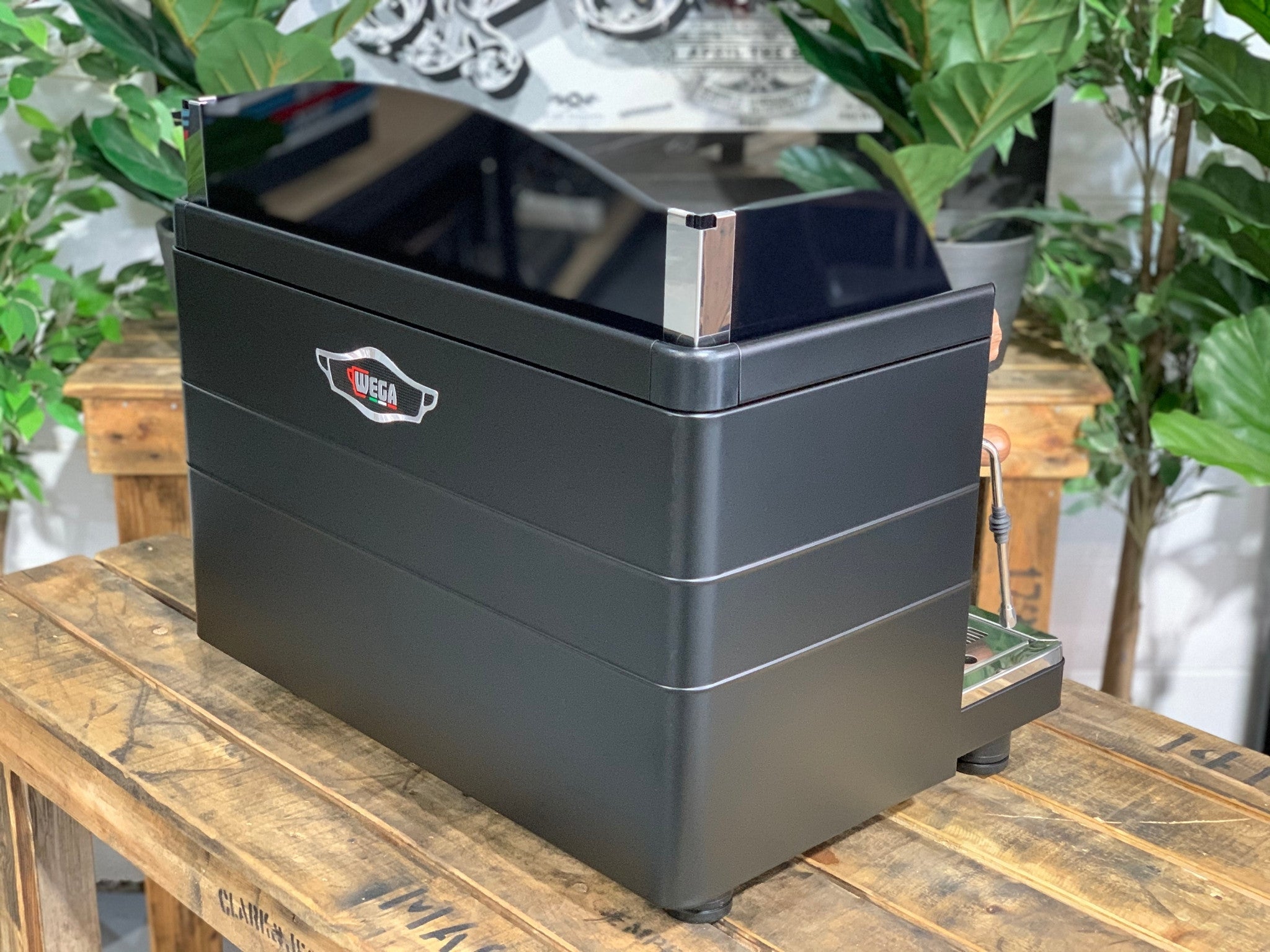 A brand new Wega Mini Nova Classic 2 Group Coffee Machine with a Matte Black finish & Timber Accents sitting on a wooden bench. There is greenery in the background. The image is taken from behind the machine and the machine is sitting on an angle so that you can see the back of the machine and the side.