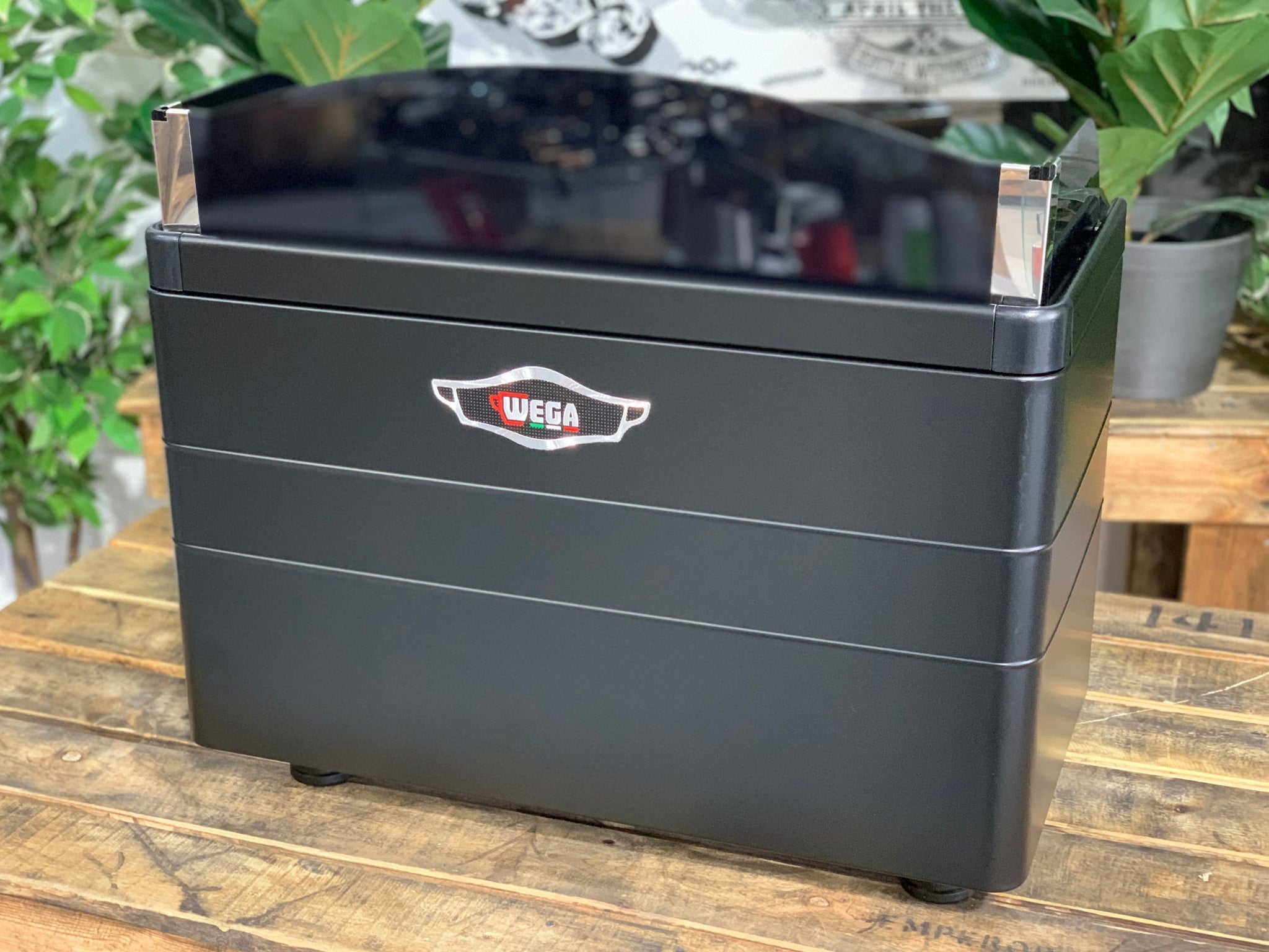 A brand new Wega Mini Nova Classic 2 Group Coffee Machine with a Matte Black finish & Timber Accents is sitting on a wooden bench. There is greenery in the background. The image is taken from behind the machine so the back is visible.
