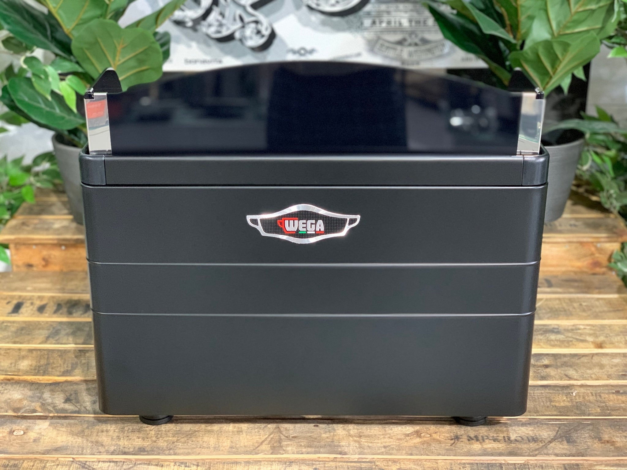 A brand new Wega Mini Nova Classic 2 Group Coffee Machine with a Matte Black finish & Timber Accents is sitting on a wooden bench. There is greenery in the background. The image is taken from behind the machine so the back is visible. The back panels of the machine are black and the Wega Logo is situated in the middle of the machine closer to the top.
