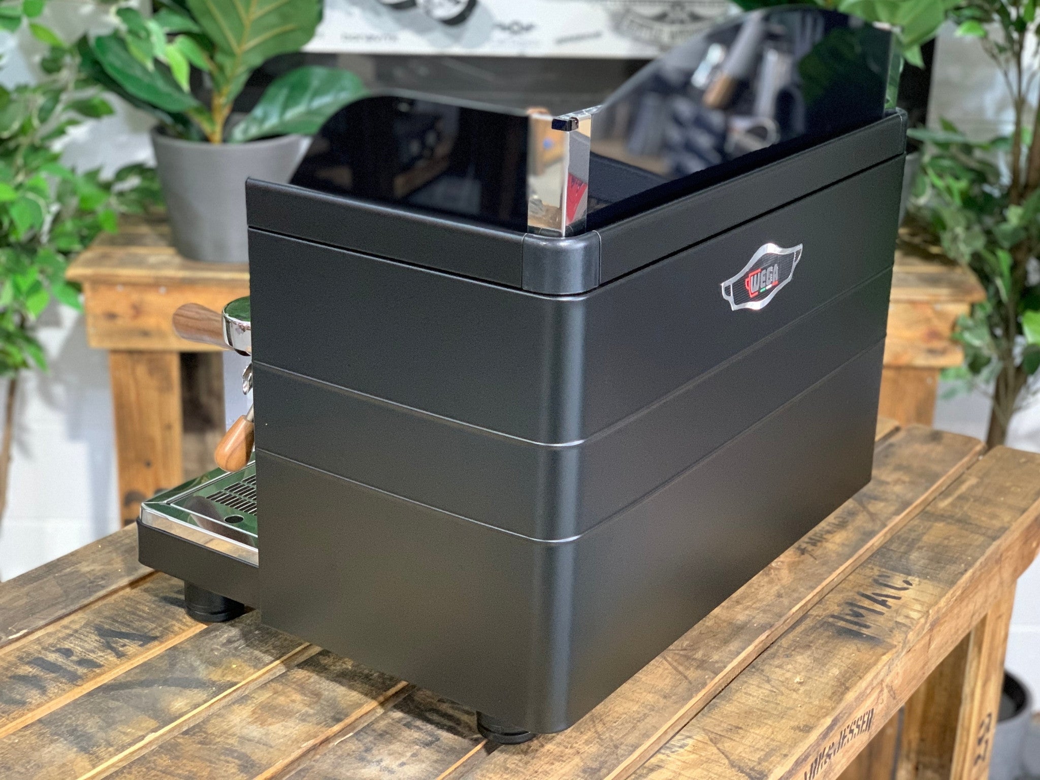 A brand new Wega Mini Nova Classic 2 Group Coffee Machine with a Matte Black finish & Timber Accents sitting on a wooden bench. There is greenery in the background. The image is taken from behind the machine and the machine is sitting on an angle so that you can see the back of the machine and the side The back panels of the machine are black and the Wega logo is situated in the middle of the machine closer to the top.