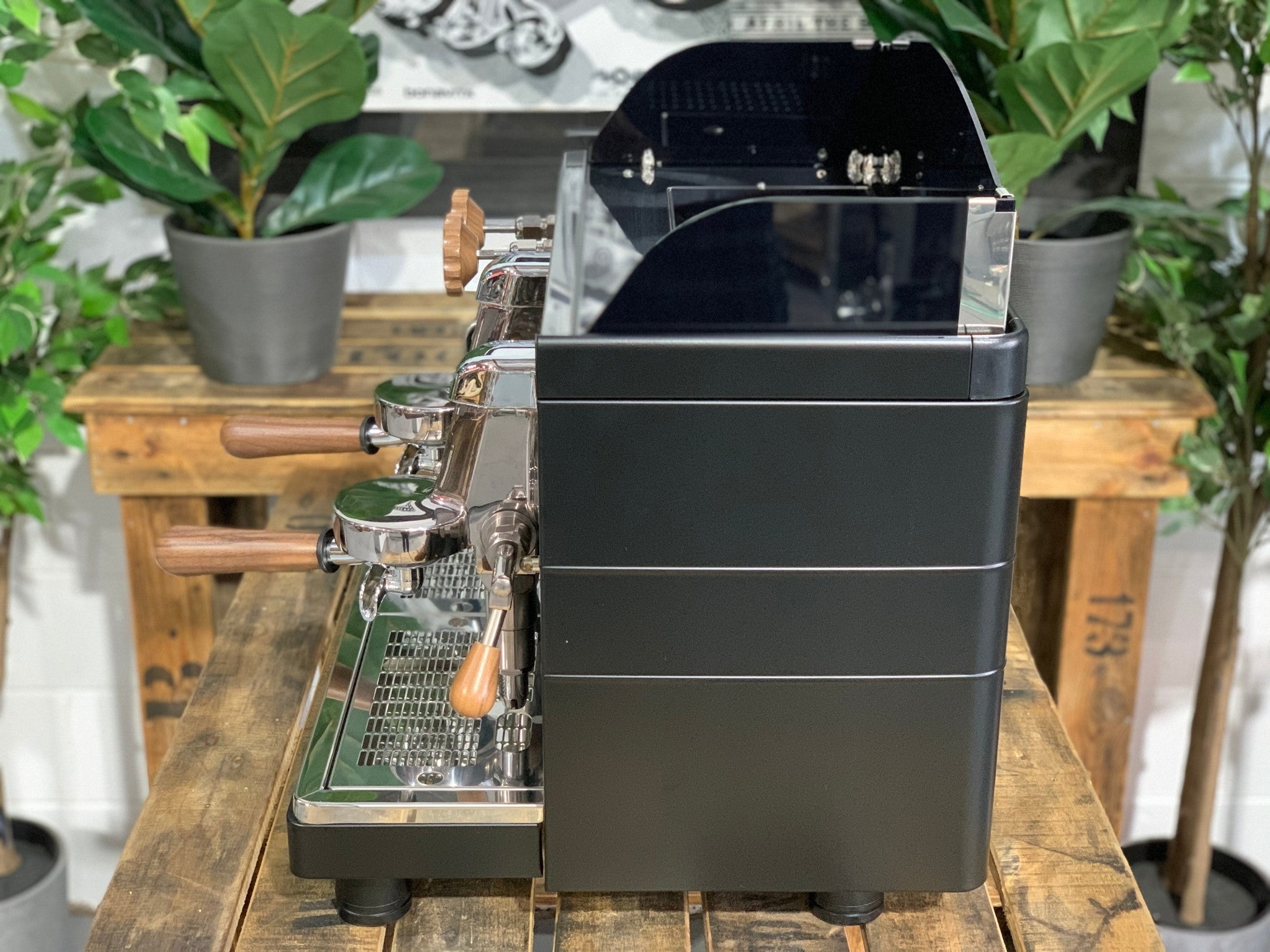 A brand new Wega Mini Nova Classic 2 Group Coffee Machine with a Matte Black finish & Timber Accents  sitting on a wooden bench. There is greenery in the background. The image is taken from the side of the machine.
