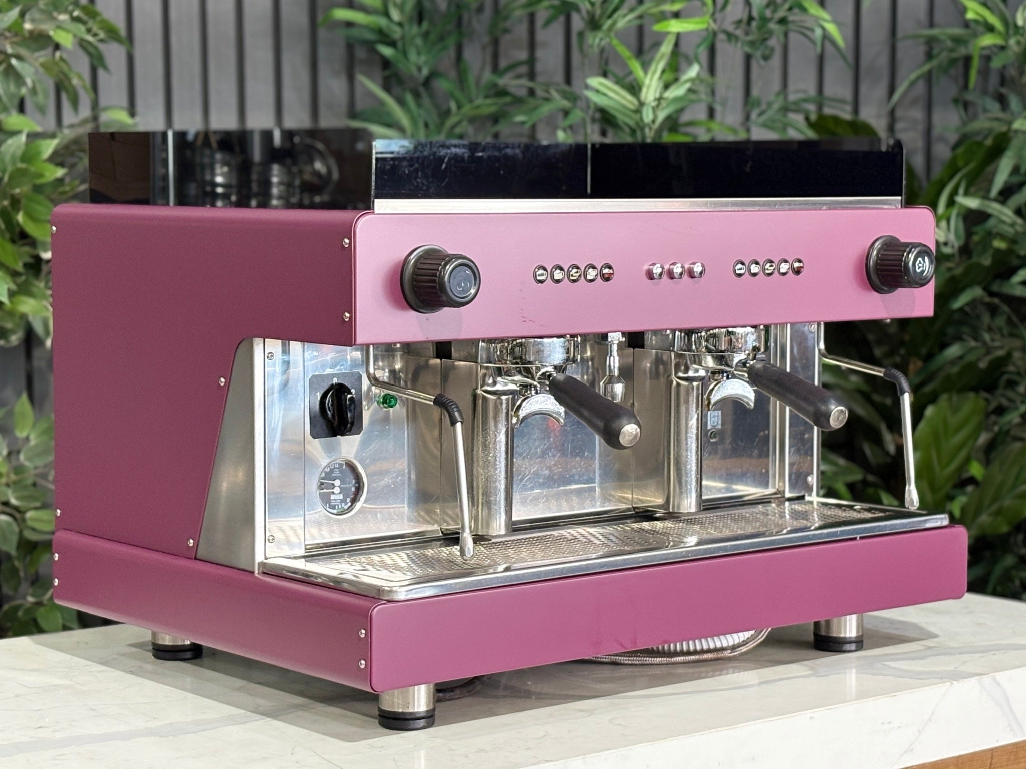 Wega Pegaso EVD 2 Group Coffee Machine Deep Pink – Used espresso coffee machine angled view showing E61 group heads, dual steam wands, and cup tray, deep pink stainless body, black acrylic top shroud, and stainless steel back panel, on a marble bench with greenery.
