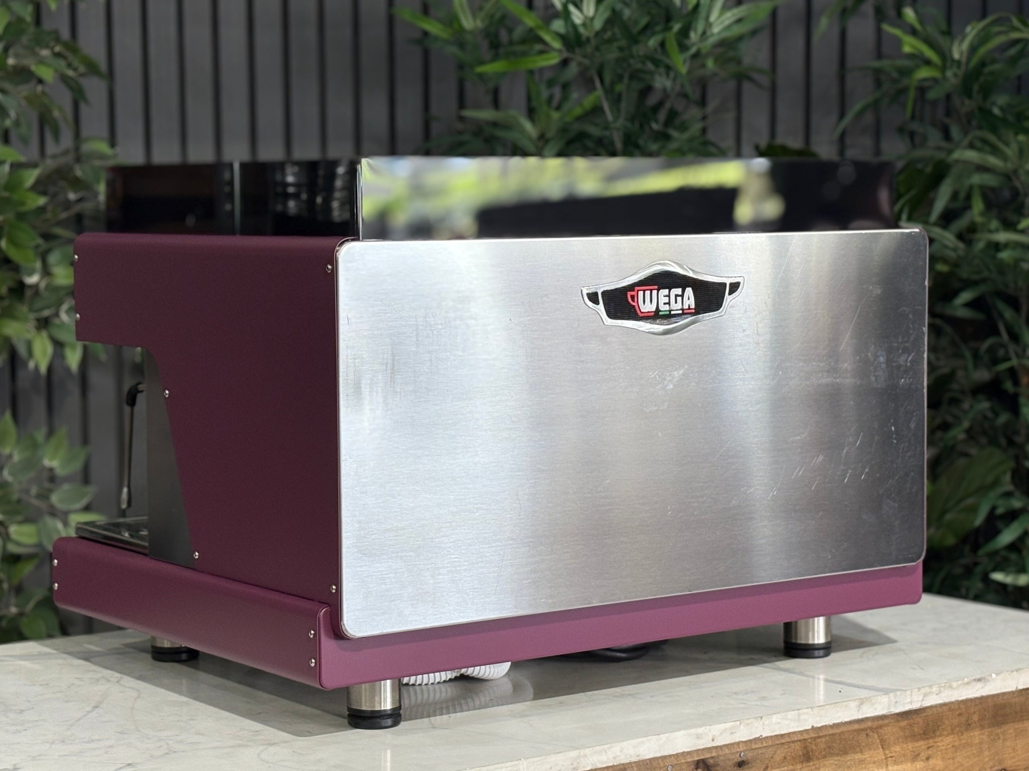 Wega Pegaso EVD 2 Group Coffee Machine Deep Pink – Used espresso coffee machine angled rear view highlighting stainless steel back panel, black acrylic top shroud, and deep pink stainless sides, placed on a marble bench with greenery.