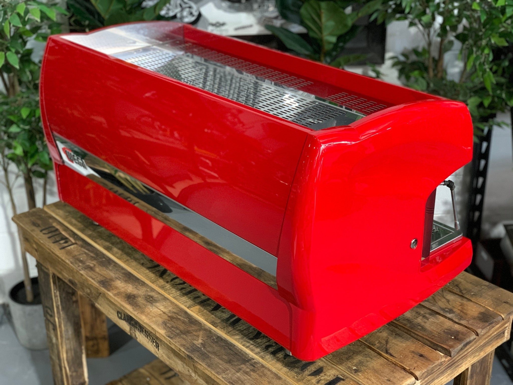 A Wega Polaris 3 Group Espresso Coffee Machine High Cup with red finish sitting on a wooden bench. There is greenery in the background. The image is taken from behind the machine and the machine is sitting on an angle so that you can see the back of the machine and the side.