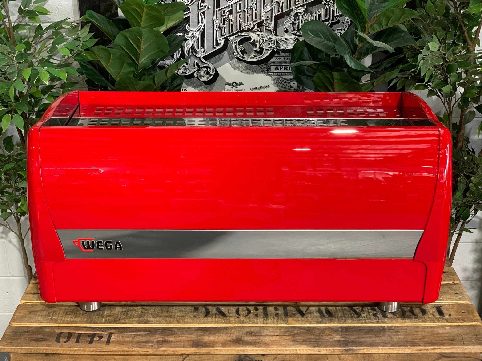 A Wega Polaris 3 Group Espresso Coffee Machine High Cup with red finish is sitting on a wooden bench. There is greenery in the background. The image is taken from behind the machine so the back is visible. The back of the machine is red, there is a thin stainless steel panel running along the back of the machine about a third down where the Wega logo is located. The logo is situated in the left hand side of that panel.