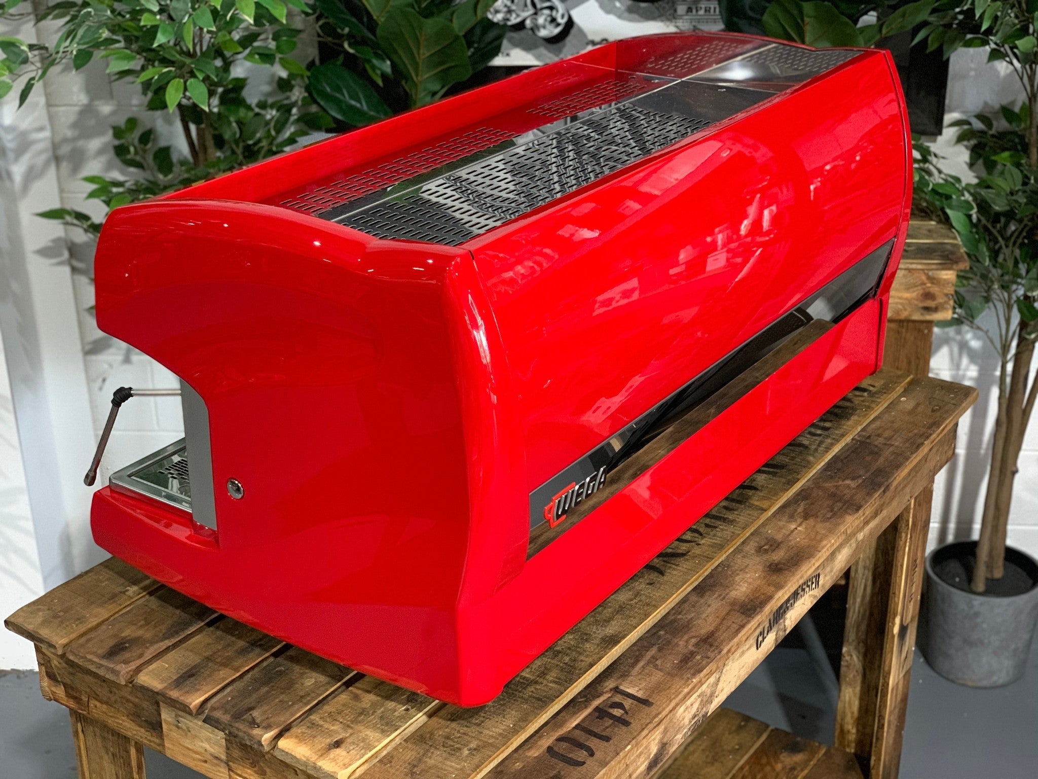 A Wega Polaris 3 Group Espresso Coffee Machine High Cup with red finish sitting on a wooden bench. There is greenery in the background. The image is taken from behind the machine and the machine is sitting on an angle so that you can see the back of the machine and the side.
