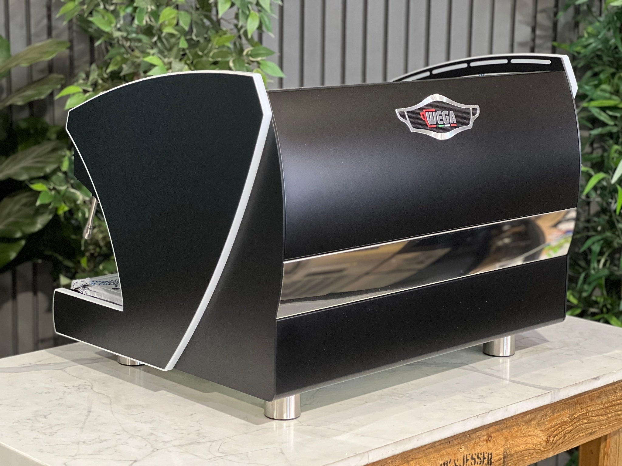 A new Wega Polaris Tron 2 Group Coffee Machine with black finish sitting on a marble bench. There is greenery in the background. The image is taken from behind the machine and the machine is sitting on an angle so that you can see the back of the machine and the side. The Wega logo is situated in the on the top of the back panel in the middle. 