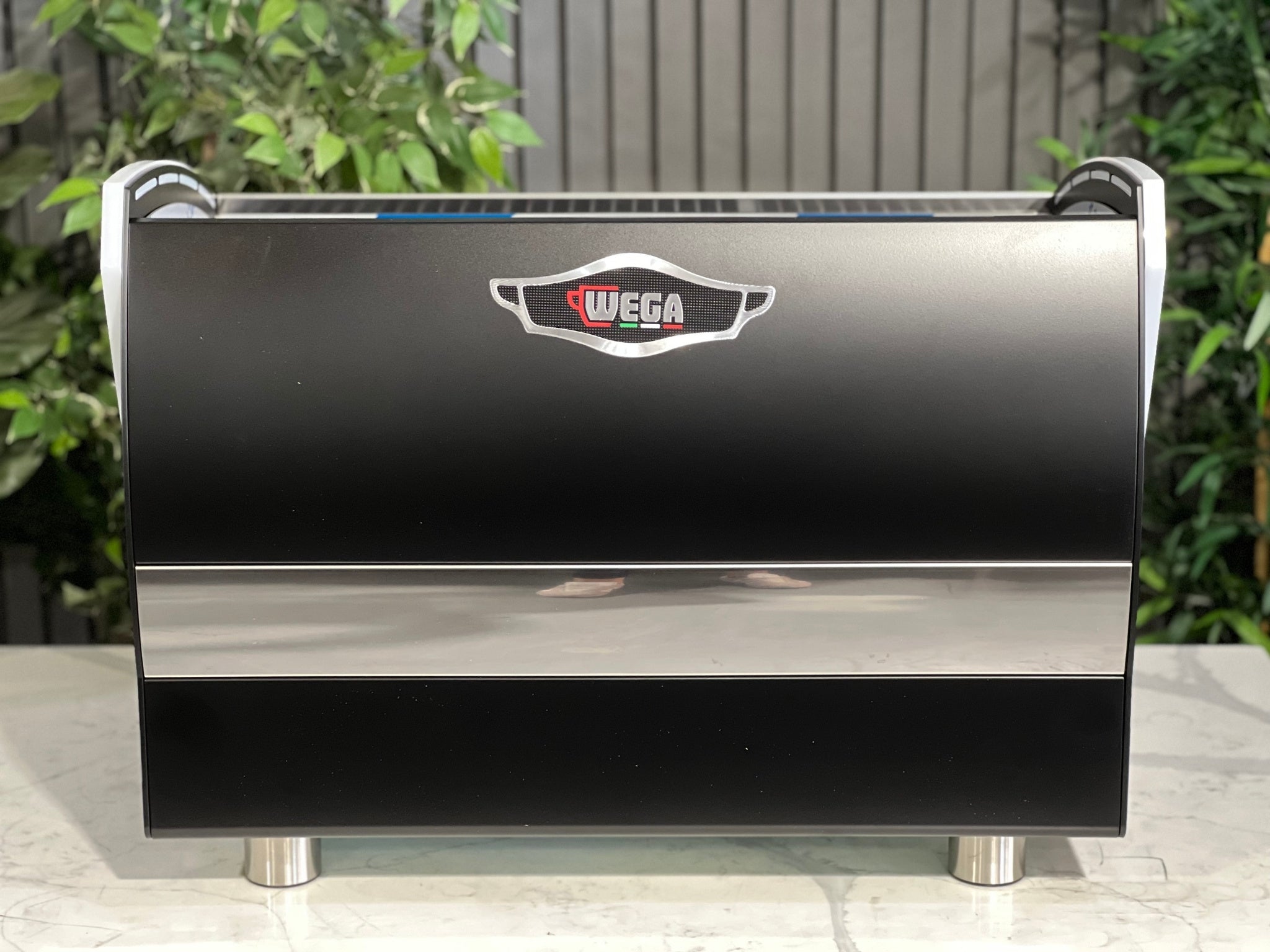A new Wega Polaris Tron 2 Group Coffee Machine with black finish  is sitting on a marble bench. There is greenery in the background. The image is taken from behind the machine so the back is visible. The Wega logo is situated in the on the top of the back panel in the middle. 
