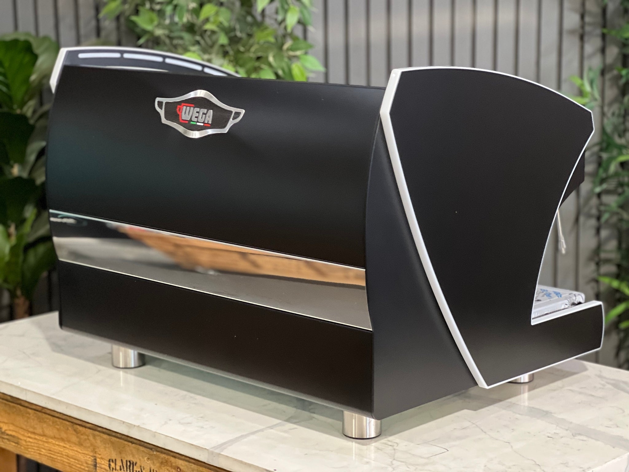 A new Wega Polaris Tron 2 Group Coffee Machine with black finish sitting on a marble bench. There is greenery in the background. The image is taken from behind the machine and the machine is sitting on an angle so that you can see the back of the machine and the side. The Wega logo is situated in the on the top of the back panel in the middle. 
