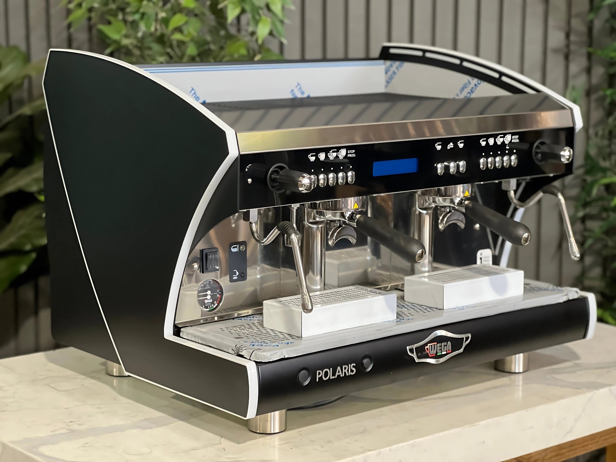 A new Wega Polaris Tron 2 Group Coffee Machine with black finish is sitting on a marble bench. There is greenery in the background. The image is taken from the front of the machine and the machine is sitting on an angle so that you can see the front of the machine and the side.
