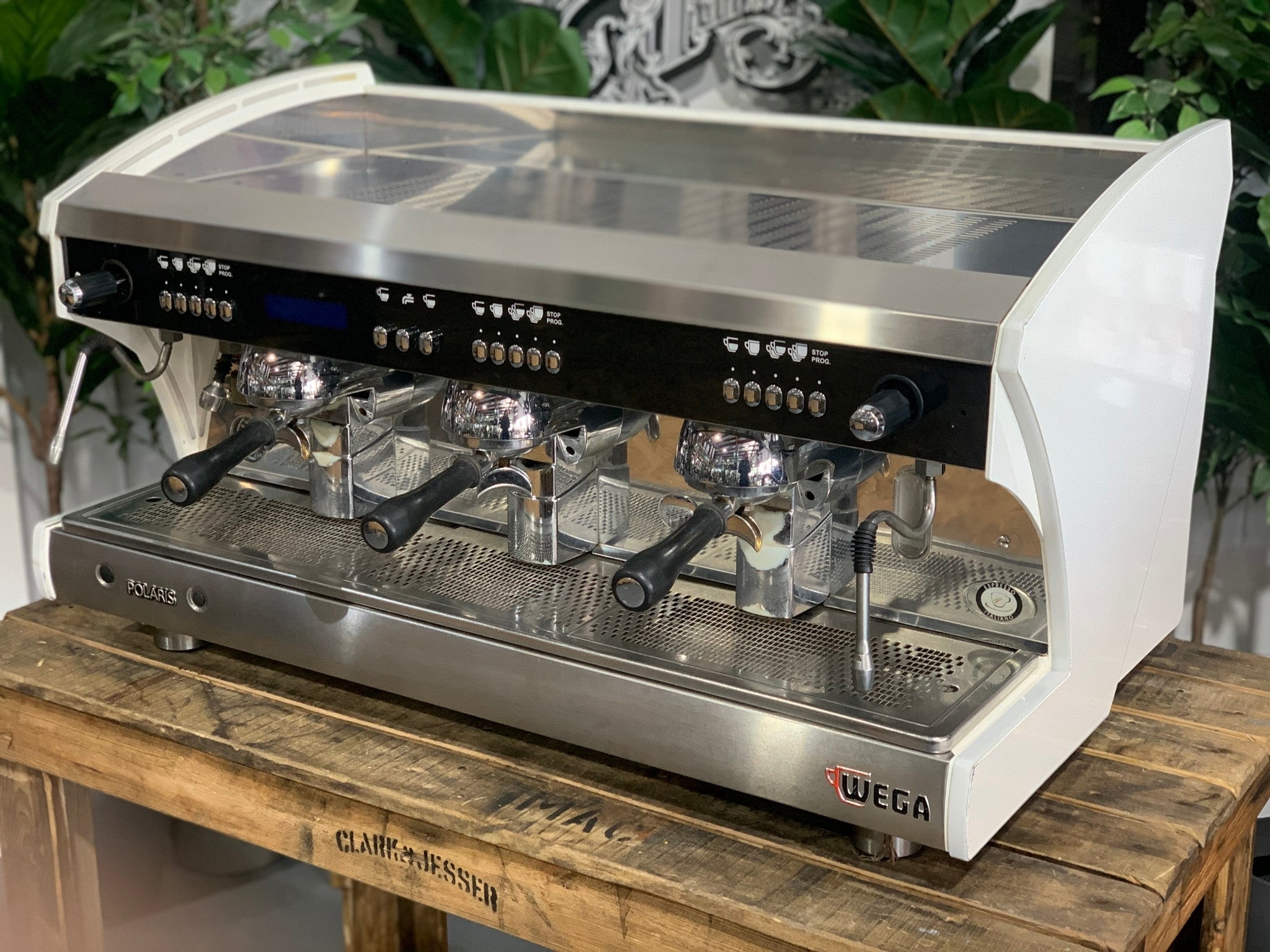A Wega Polaris Tron 3 Group Espresso Coffee Machine with white finish  is sitting on a wooden bench. There is greenery in the background. The image is taken from the front of the machine and the machine is sitting on an angle so that you can see the front of the machine and the side.