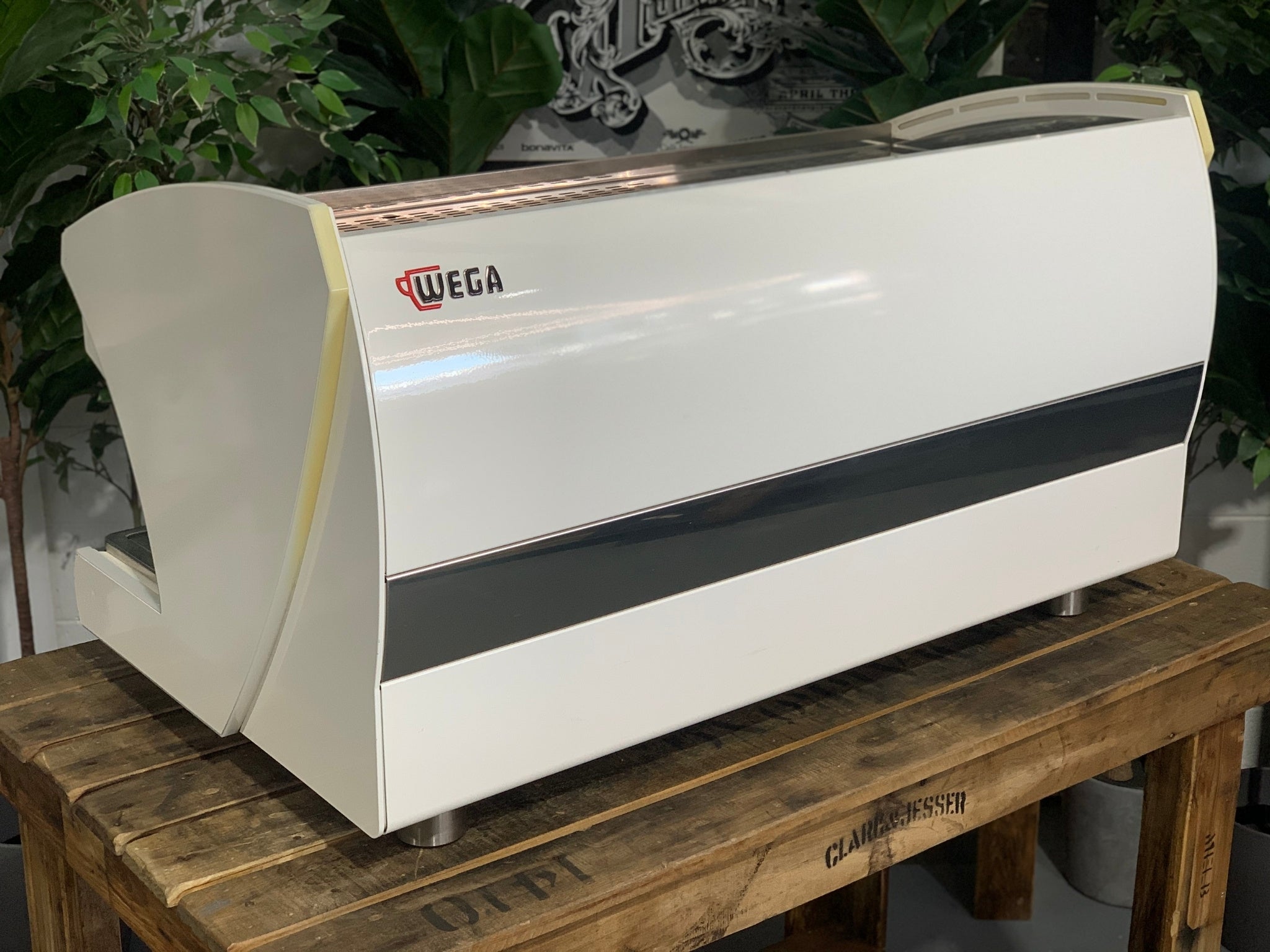 A Wega Polaris Tron 3 Group Espresso Coffee Machine with white finish sitting on a wooden bench. There is greenery in the background. The image is taken from behind the machine and the machine is sitting on an angle so that you can see the back of the machine and the side.