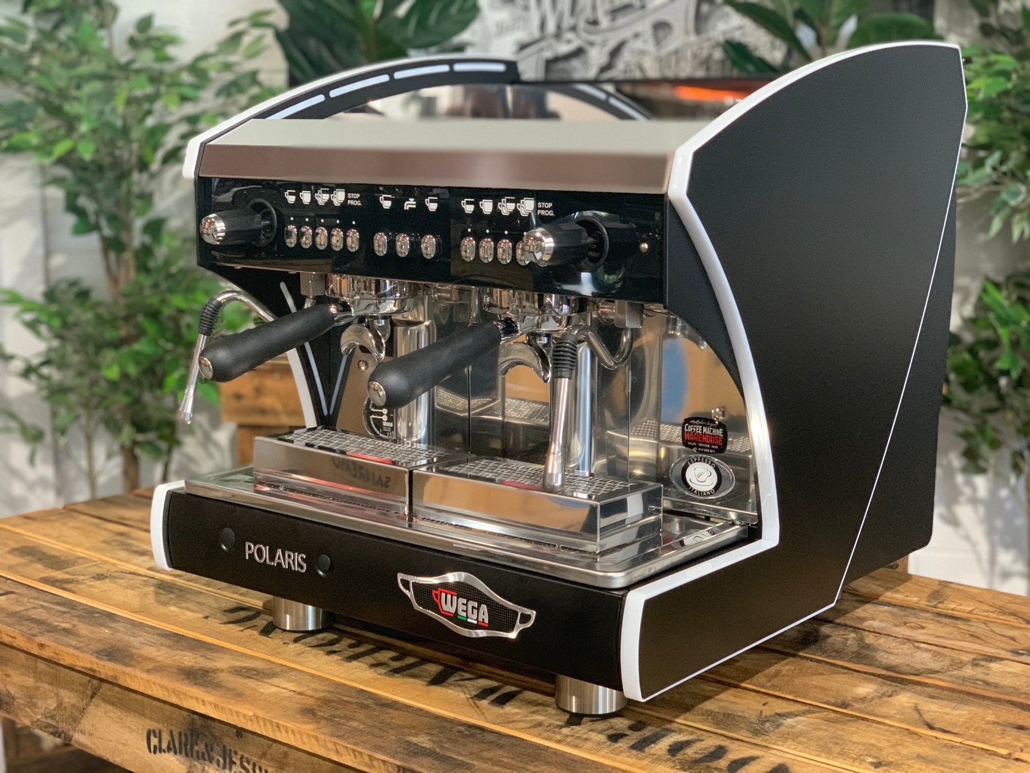 A new Wega Polaris Tron Compact 2 Group Espresso Coffee Machine with black finish  is sitting on a wooden bench. There is greenery in the background. The image is taken from the front of the machine and the machine is sitting on an angle so that you can see the front of the machine and the side.