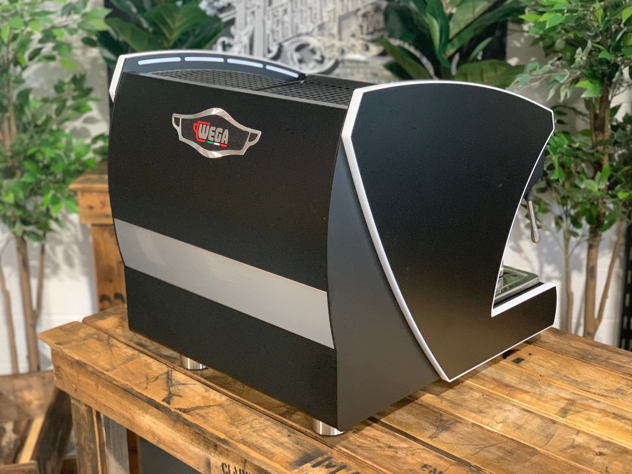 A new Wega Polaris Tron Compact 2 Group Espresso Coffee Machine with black finish sitting on a wooden bench. There is greenery in the background. The image is taken from behind the machine and the machine is sitting on an angle so that you can see the back of the machine and the side.
The Wega logo is situated in the middle close to the top of the back panel of the machine. 