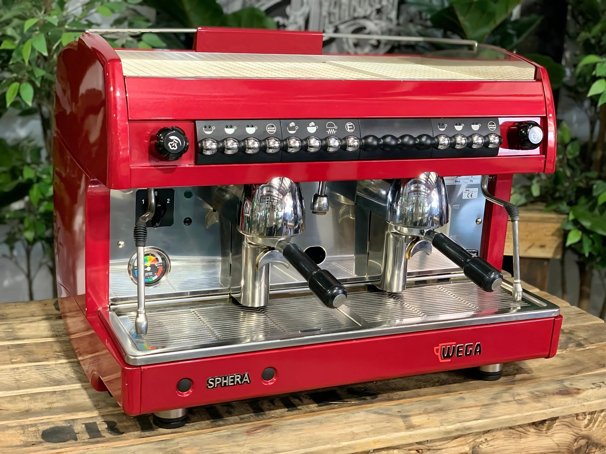 A Wega Sphera 2 Group Espresso Coffee Machine with Red finish is sitting on a wooden bench. There is greenery in the background. The image is taken from the front of the machine and the machine is sitting on an angle so that you can see the front of the machine and the side.