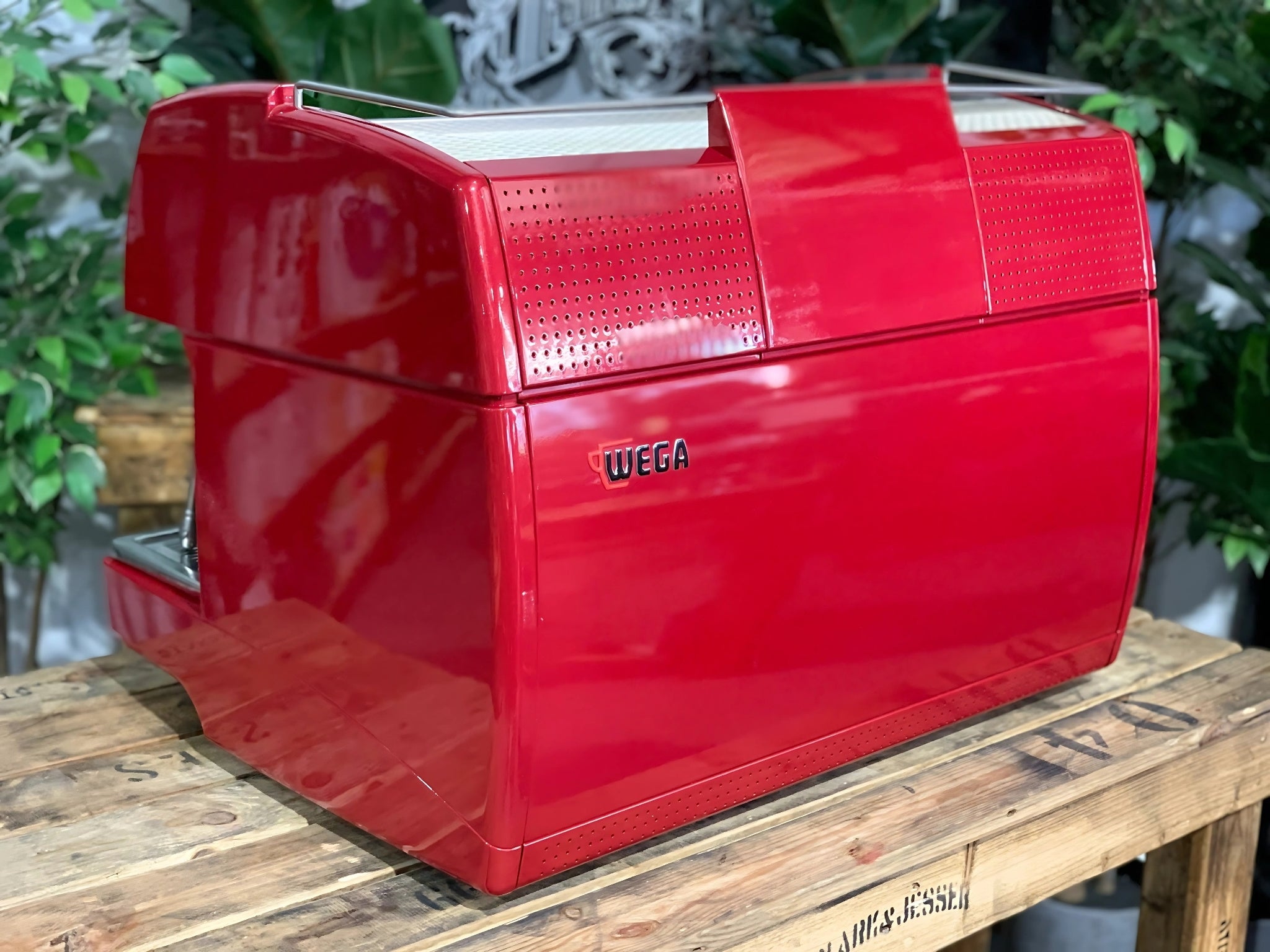 A Wega Sphera 2 Group Espresso Coffee Machine with Red finishsitting on a wooden bench. There is greenery in the background. The image is taken from behind the machine and the machine is sitting on an angle so that you can see the back of the machine and the side. The machine is red and the Wega logo is on the back of the machine placed in the middle of the left hand side. 