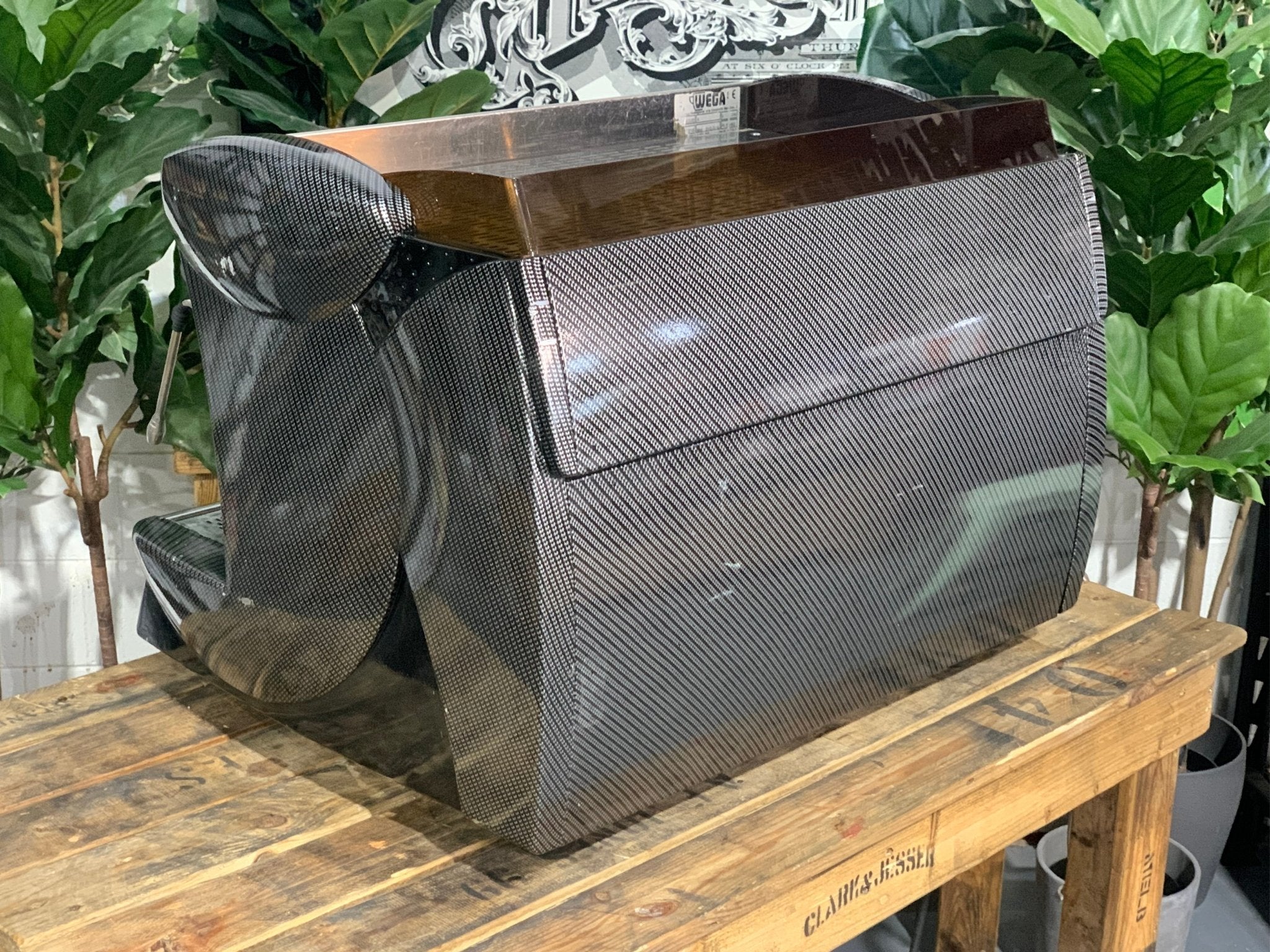 A Wega Vela 2 Group Espresso Coffee Machine with Carbon Fibre panels sitting on a wooden bench. There is greenery in the background. The image is taken from behind the machine and the machine is sitting on an angle so that you can see the back of the machine and the side.