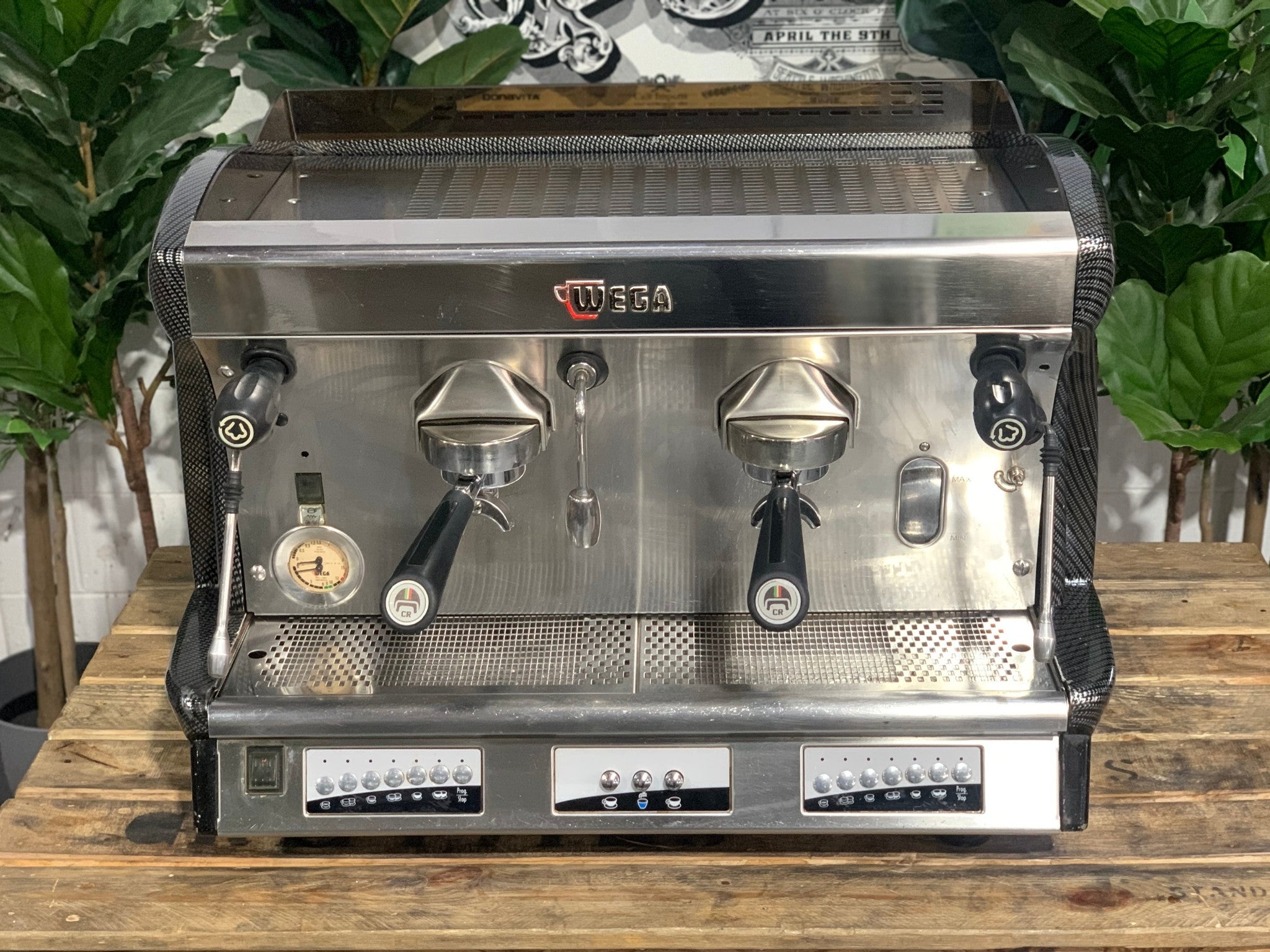 A Wega Vela 2 Group Espresso Coffee Machine with Carbon Fibre panels sitting on a wooden bench. There is greenery in the background. The image is taken from the front of the machine so that you can see the front of the machine, control panel, buttons and group heads.
