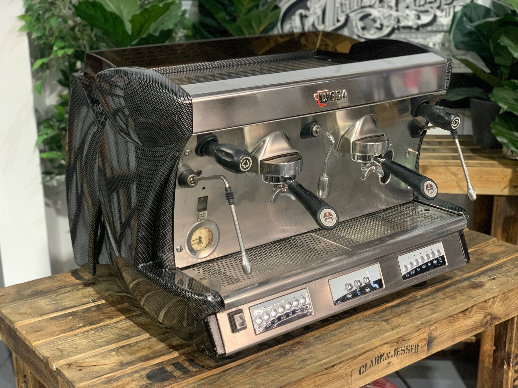A Wega Vela 2 Group Espresso Coffee Machine with Carbon Fibre panels  is sitting on a wooden bench. There is greenery in the background. The image is taken from the front of the machine and the machine is sitting on an angle so that you can see the front of the machine and the side.
