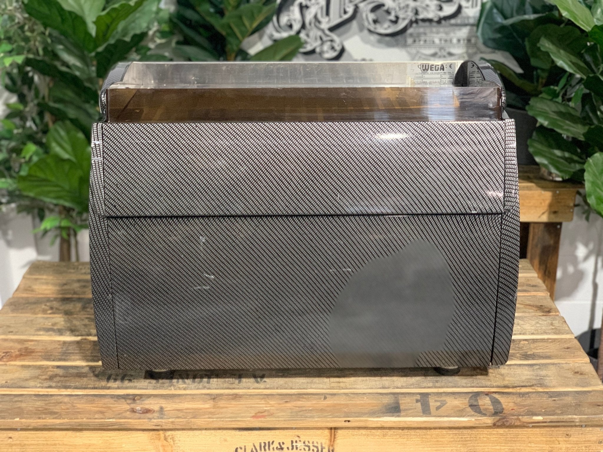 A Wega Vela 2 Group Espresso Coffee Machine with Carbon Fibre panels 
is sitting on a wooden bench. There is greenery in the background. The image is taken from behind the machine so the back is visible.
