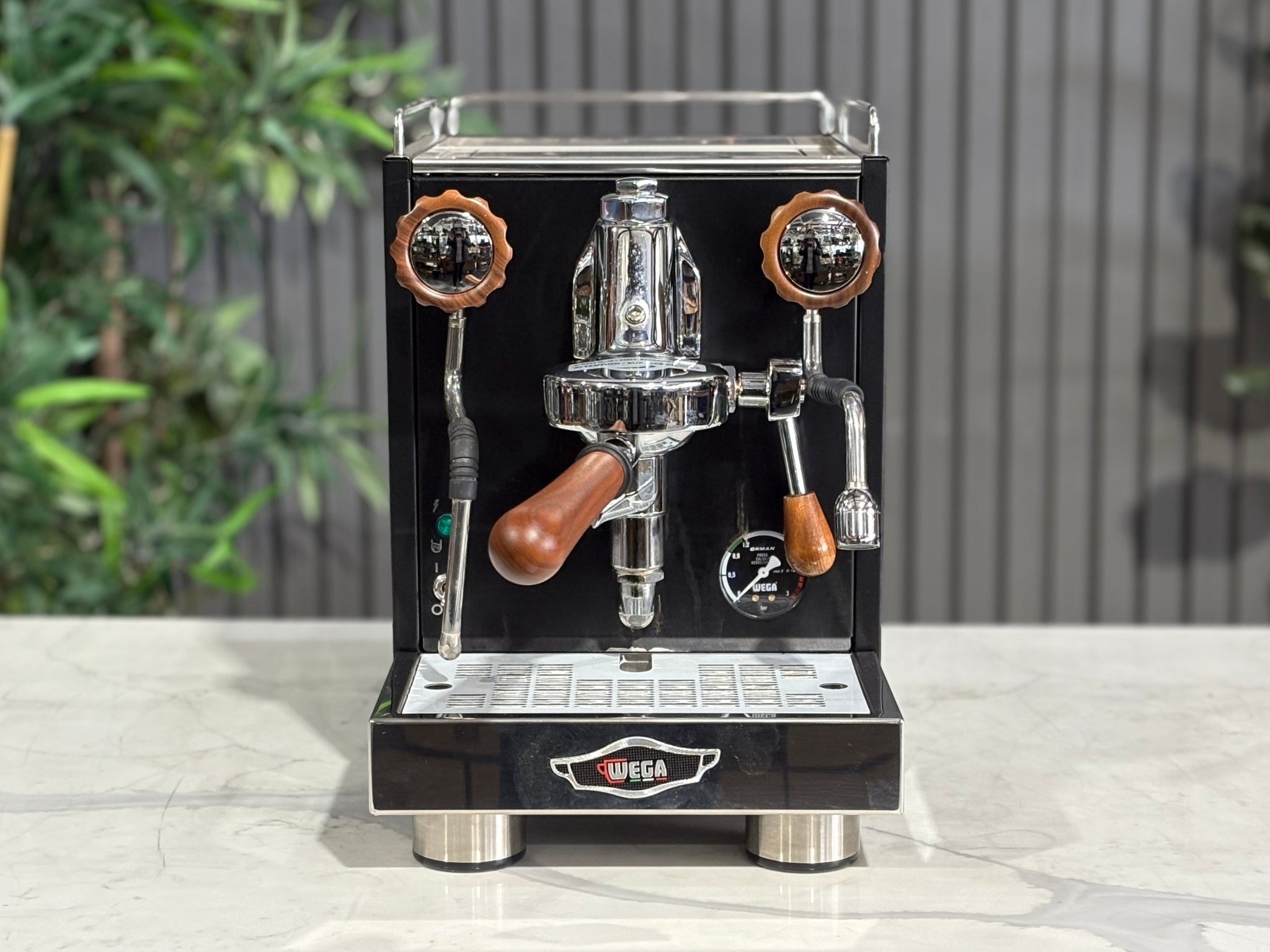 A Wega W Mini 1 Group Coffee Machine Matte Black finish & Timber accents sitting on a marble bench. There is greenery in the background. The image is taken from the front of the machine so that you can see the front of the machine, control panel, buttons and group head.