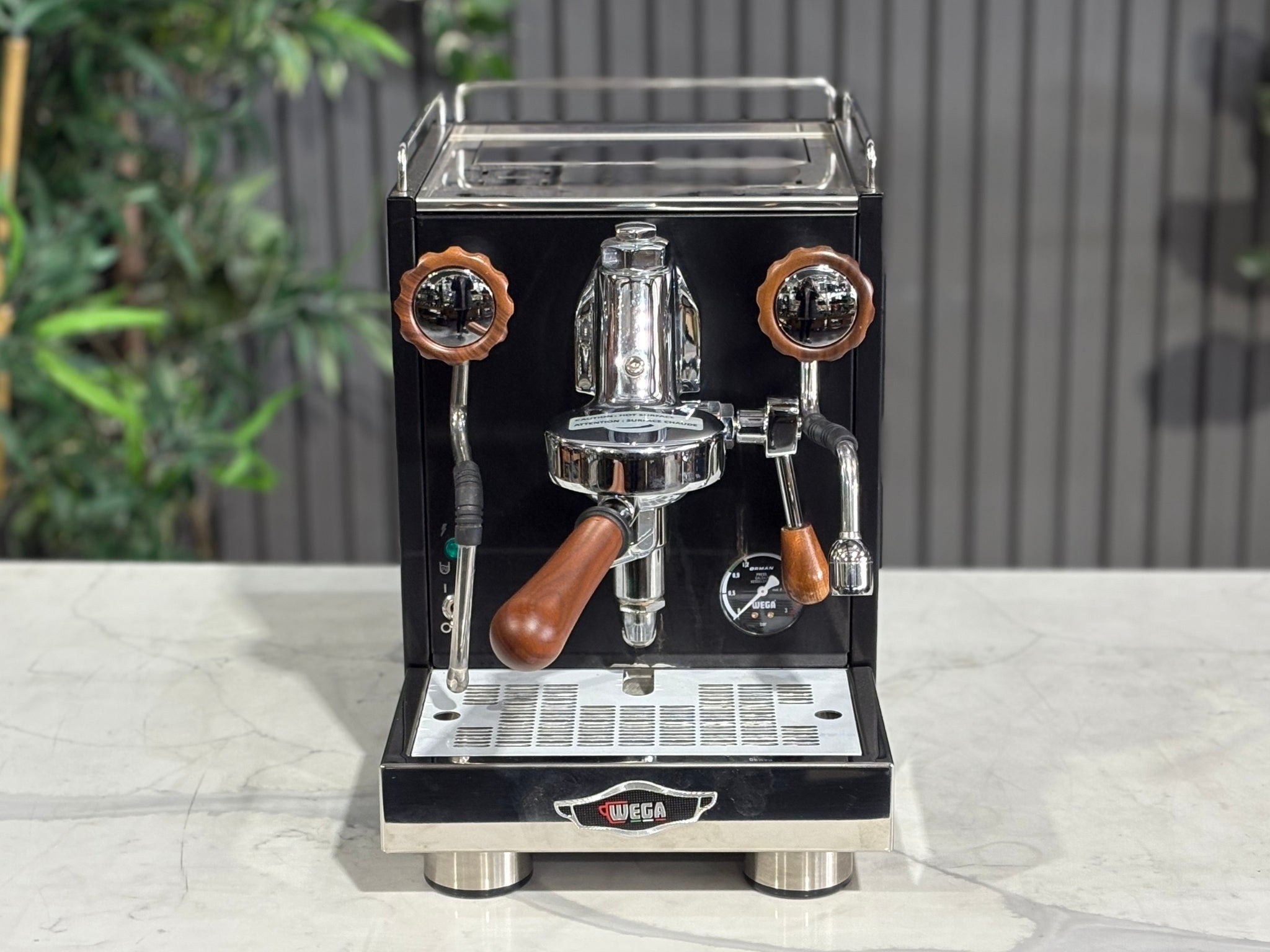 A Wega W Mini 1 Group Coffee Machine Matte Black finish & Timber accents
sitting on a marble bench. There is greenery in the background. The image is taken from the front of the machine but at a high angle so that you can see the front of the machine and also the top of the machine.
