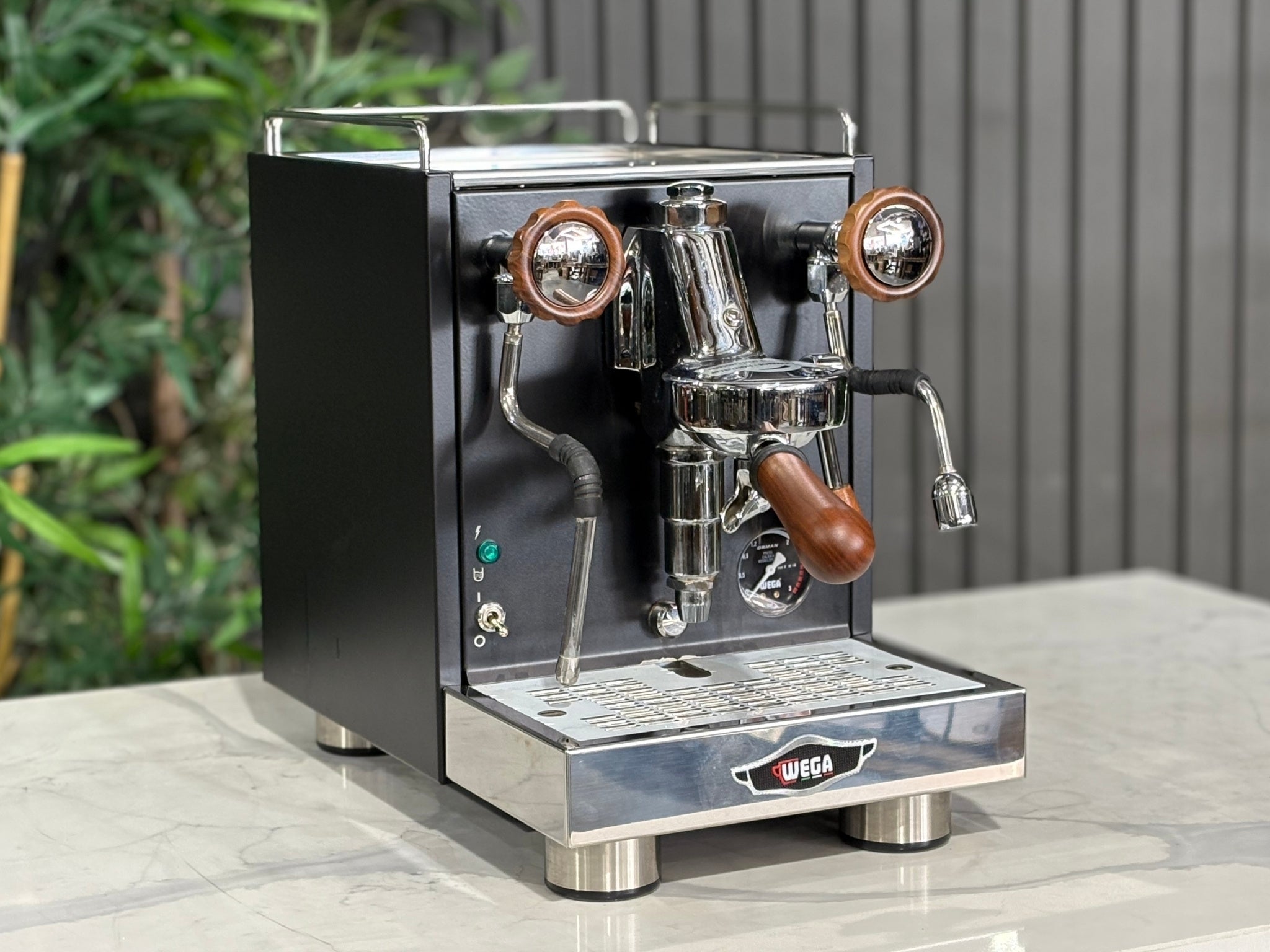A Wega W Mini 1 Group Coffee Machine Matte Black finish & Timber accents l is sitting on a marble bench. There is greenery in the background. The image is taken from the front of the machine and the machine is sitting on an angle so that you can see the front of the machine and the side. 