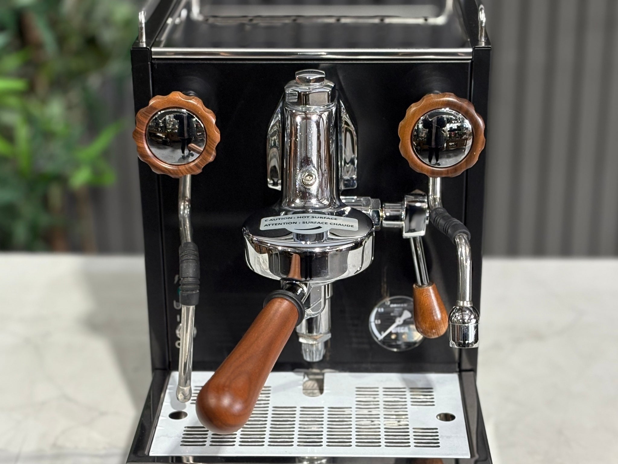 A Wega W Mini 1 Group Coffee Machine Matte Black finish & Timber accents s sitting on a marble bench. There is greenery in the background. The image is a close up image taken from the front of the machine so that you can see the front of the machine, the controls including rotary knobs, group handle and steam wand.
