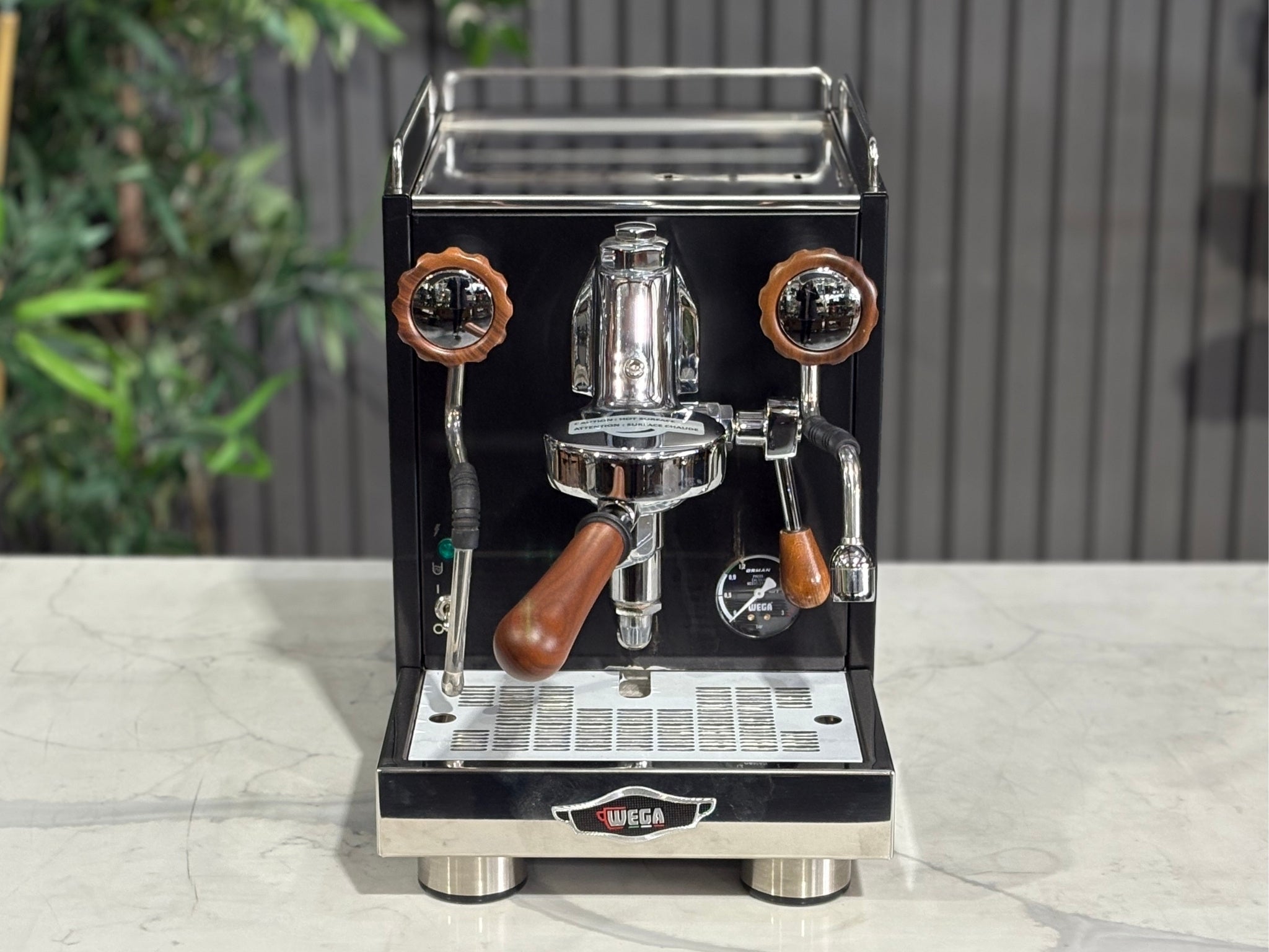 A Wega W Mini 1 Group Coffee Machine Matte Black finish & Timber accents
sitting on a marble bench. There is greenery in the background. The image is taken from the front of the machine but at a high angle so that you can see the front of the machine and also the top of the machine.