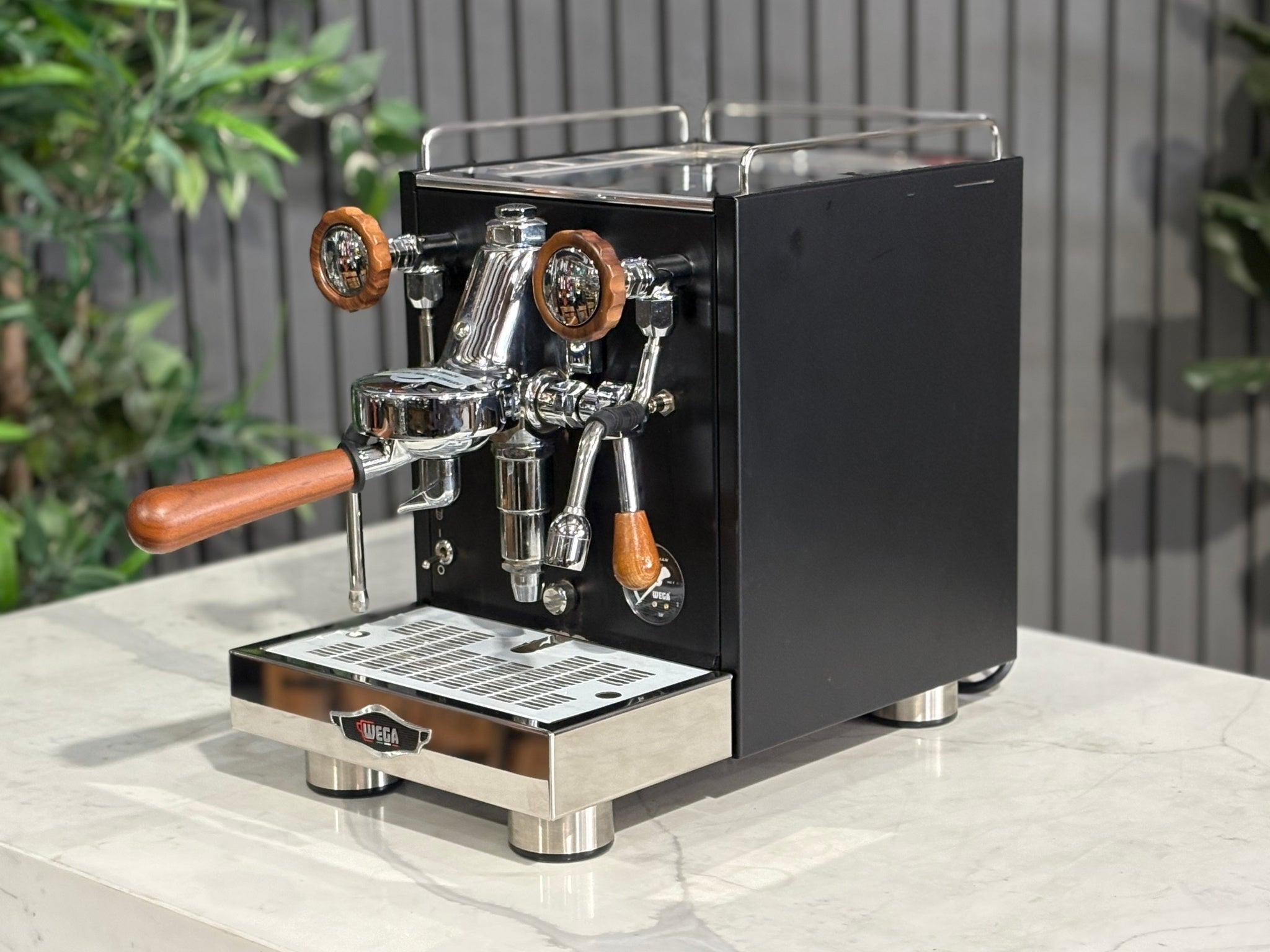A Wega W Mini 1 Group Coffee Machine Matte Black finish & Timber accents l is sitting on a marble bench. There is greenery in the background. The image is taken from the front of the machine and the machine is sitting on an angle so that you can see the front of the machine and the side. 
