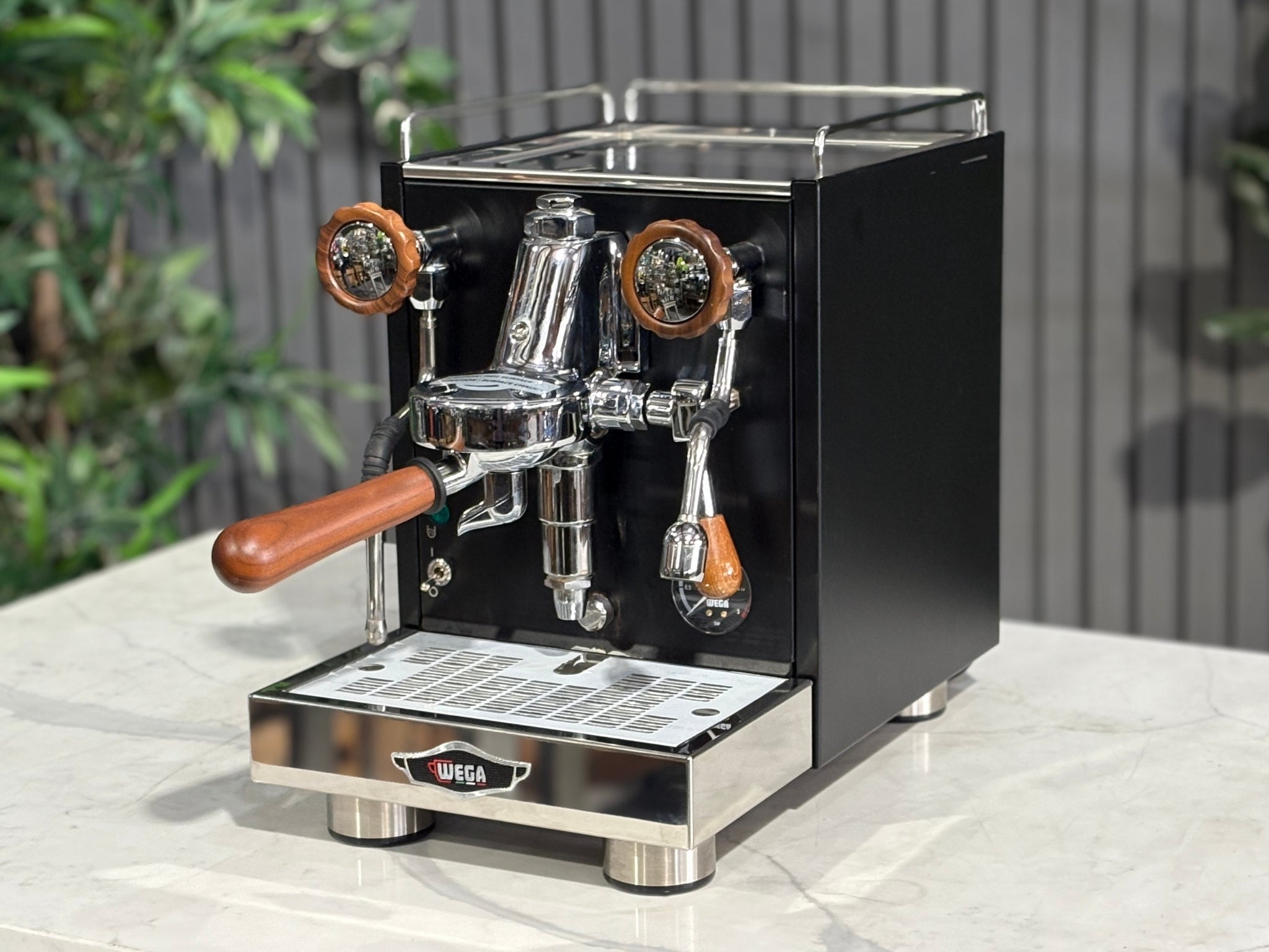 A Wega W Mini 1 Group Coffee Machine Matte Black finish & Timber accents l is sitting on a marble bench. There is greenery in the background. The image is taken from the front of the machine and the machine is sitting on an angle so that you can see the front of the machine and the side. 