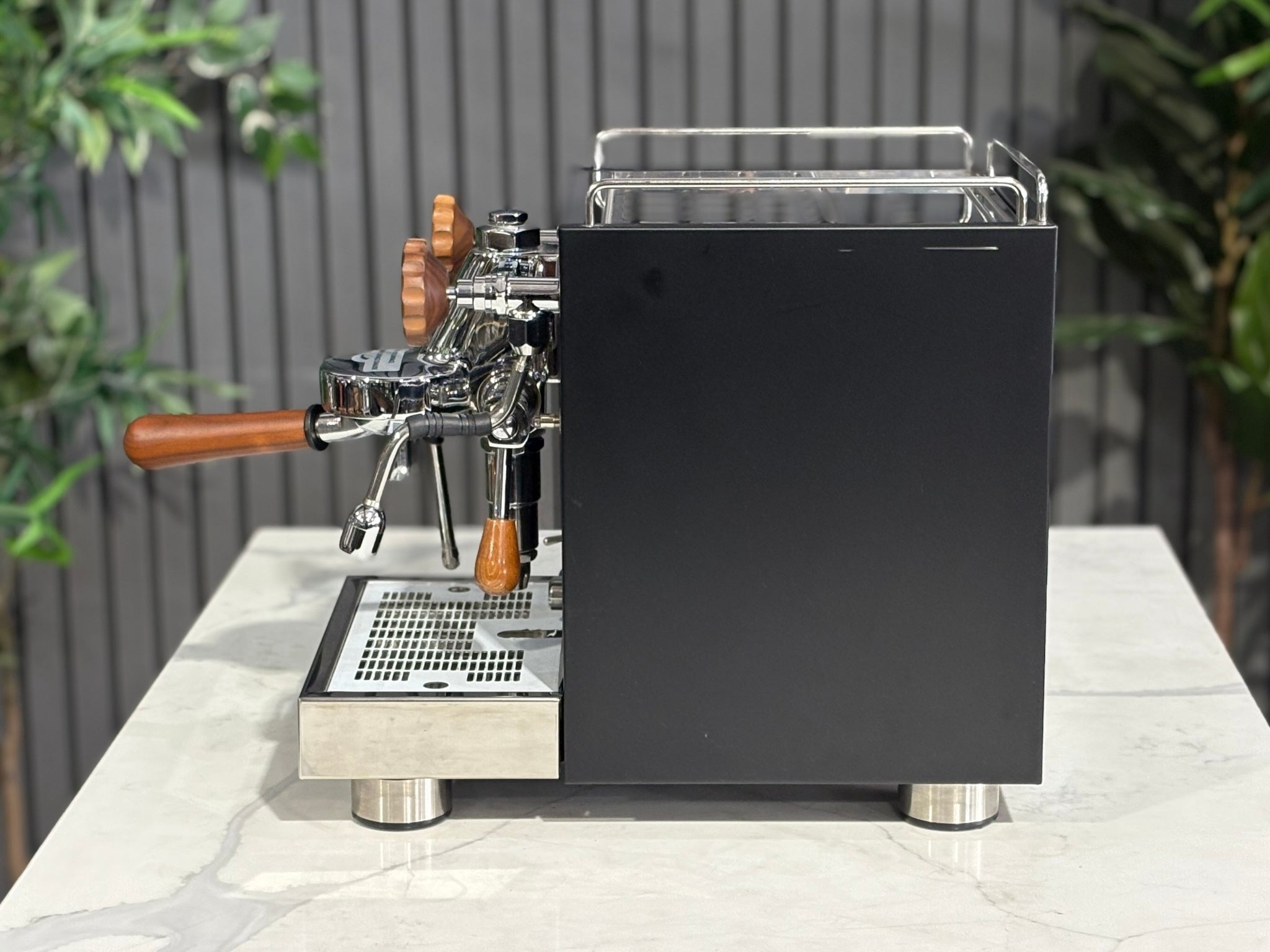 A Wega W Mini 1 Group Coffee Machine Matte Black finish & Timber accents sitting on a marble bench. There is greenery in the background. The image is taken from the side of the machine.