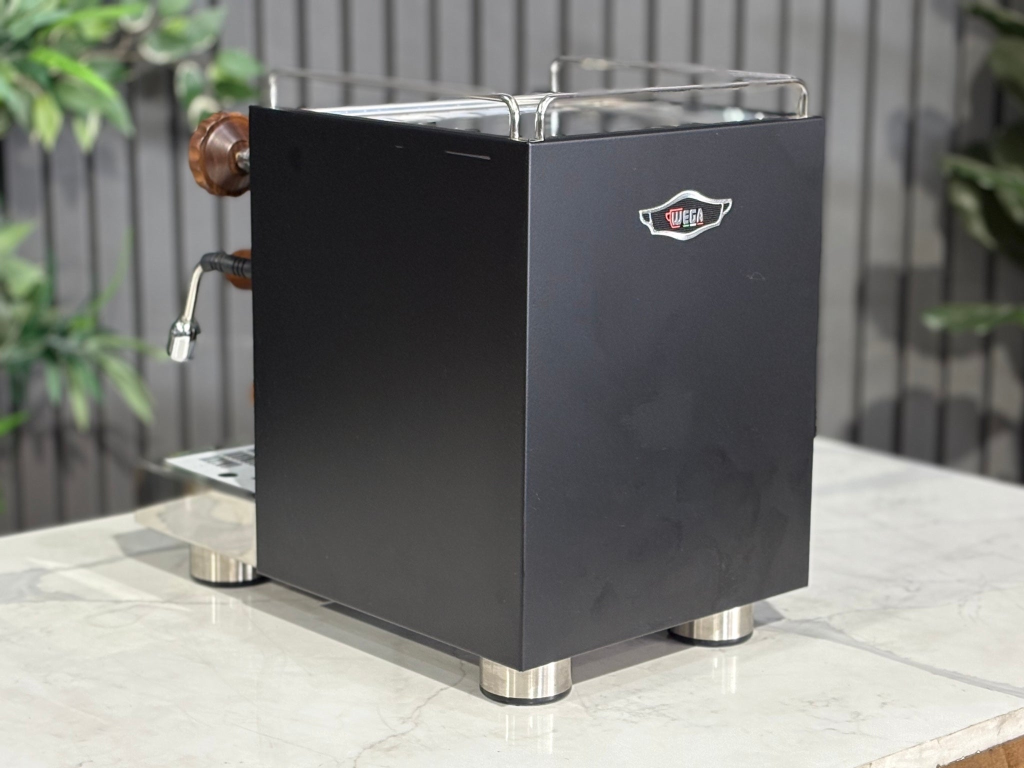 A Wega W Mini 1 Group Coffee Machine Matte Black finish & Timber accents sitting on a marble bench. There is greenery in the background. The image is taken from behind the machine and the machine is sitting on an angle so that you can see the back of the machine and the side.