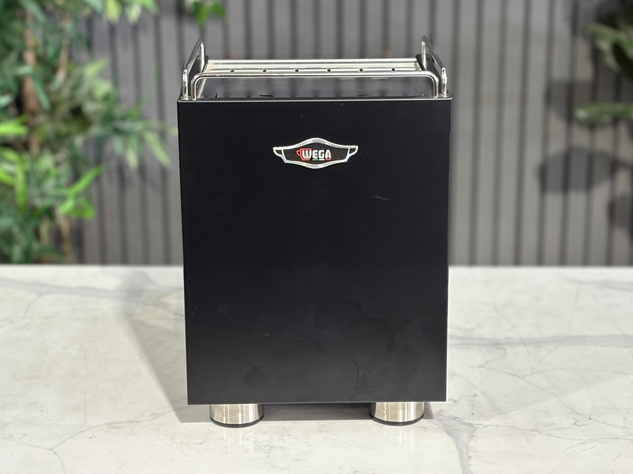 A Wega W Mini 1 Group Coffee Machine Matte Black finish & Timber accents s sitting on a marble bench. There is greenery in the background. The image is taken from behind the machine so the back is visible. The back of the machine and the Wega logo is situated in the middle at the top of the machine. 
