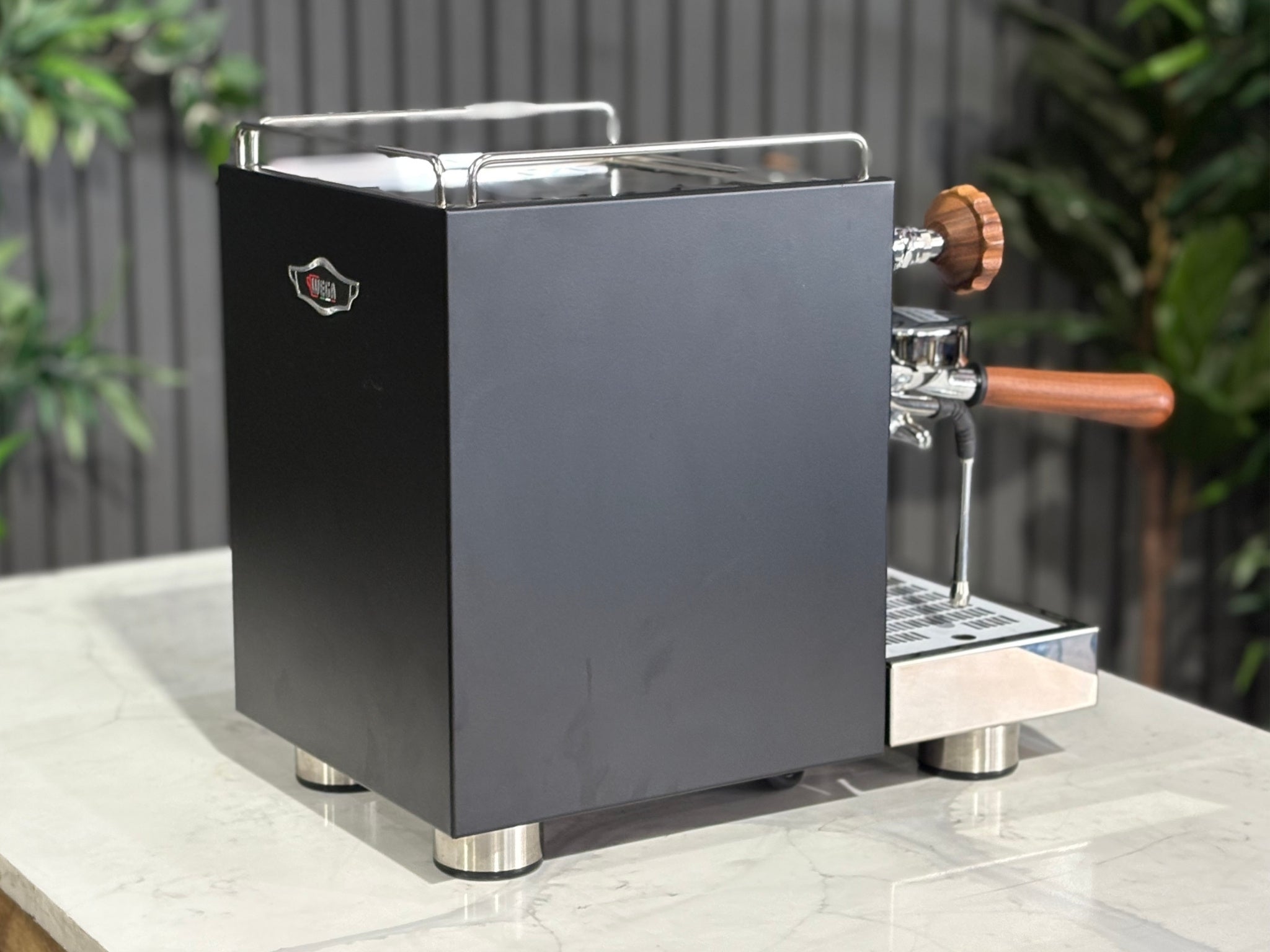 A Wega W Mini 1 Group Coffee Machine Matte Black finish & Timber accents sitting on a marble bench. There is greenery in the background. The image is taken from behind the machine and the machine is sitting on an angle so that you can see the back of the machine and the side.