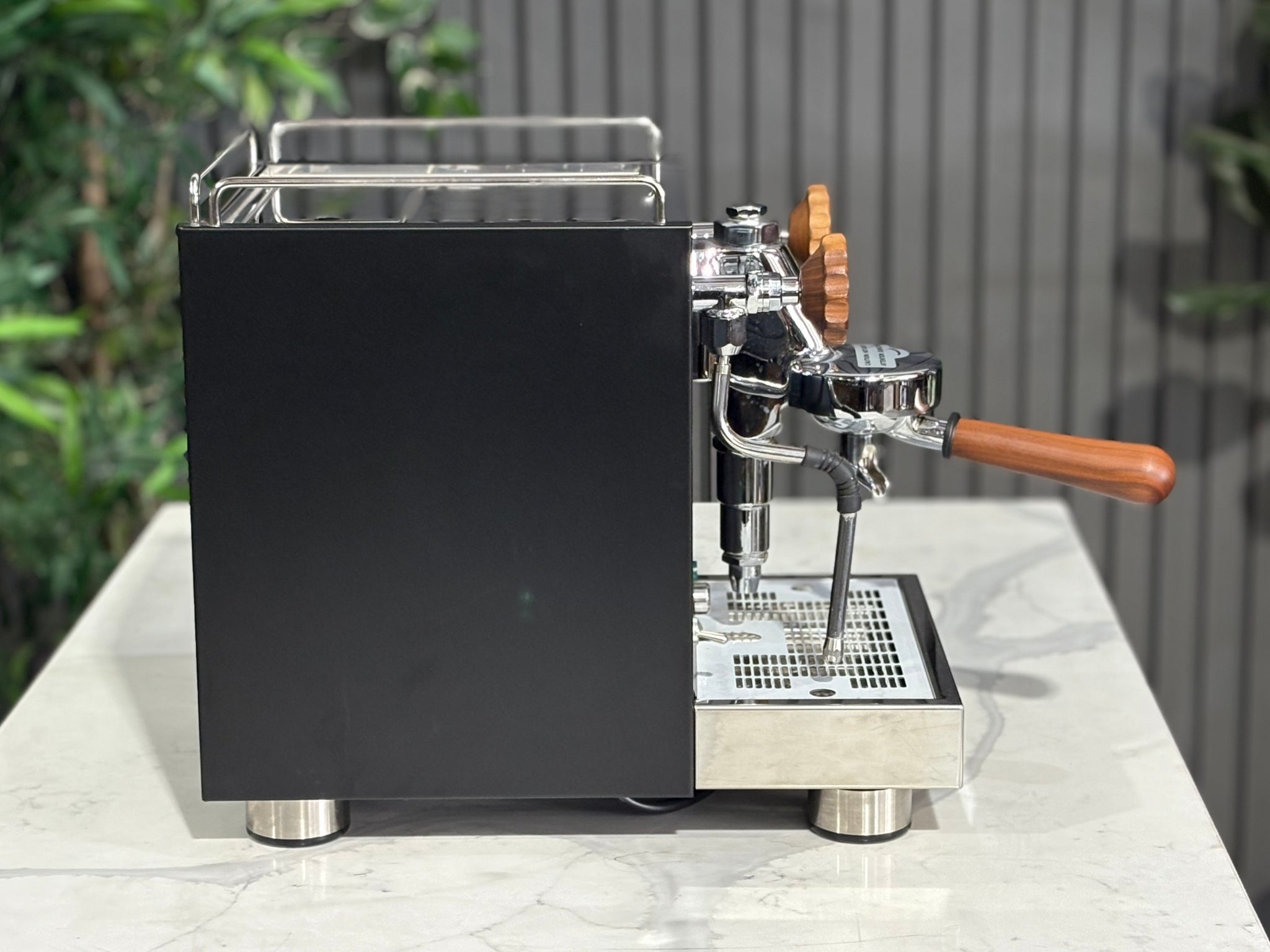 A Wega W Mini 1 Group Coffee Machine Matte Black finish & Timber accents sitting on a marble bench. There is greenery in the background. The image is taken from the side of the machine.