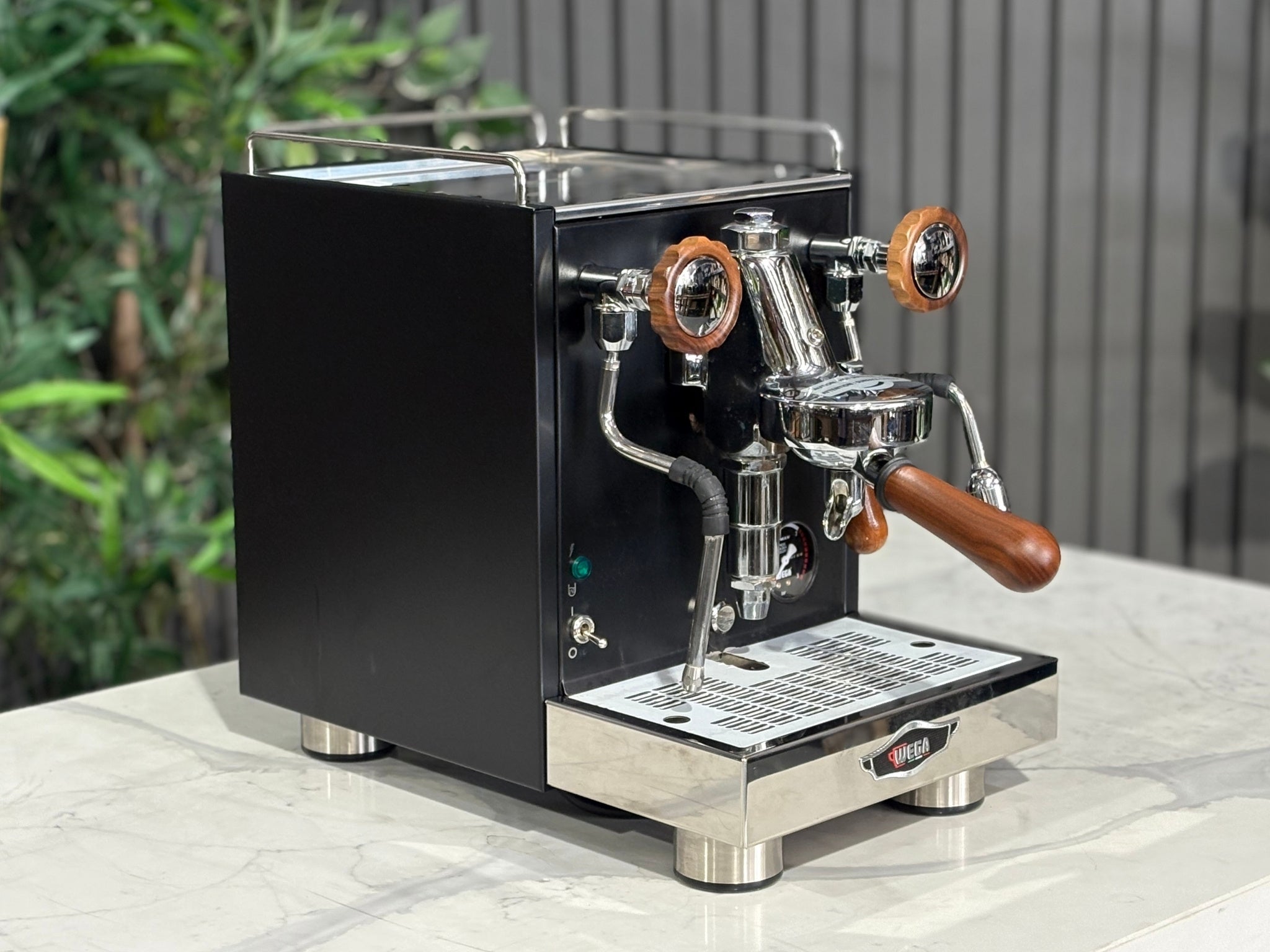 A Wega W Mini 1 Group Coffee Machine Matte Black finish & Timber accents l is sitting on a marble bench. There is greenery in the background. The image is taken from the front of the machine and the machine is sitting on an angle so that you can see the front of the machine and the side. 